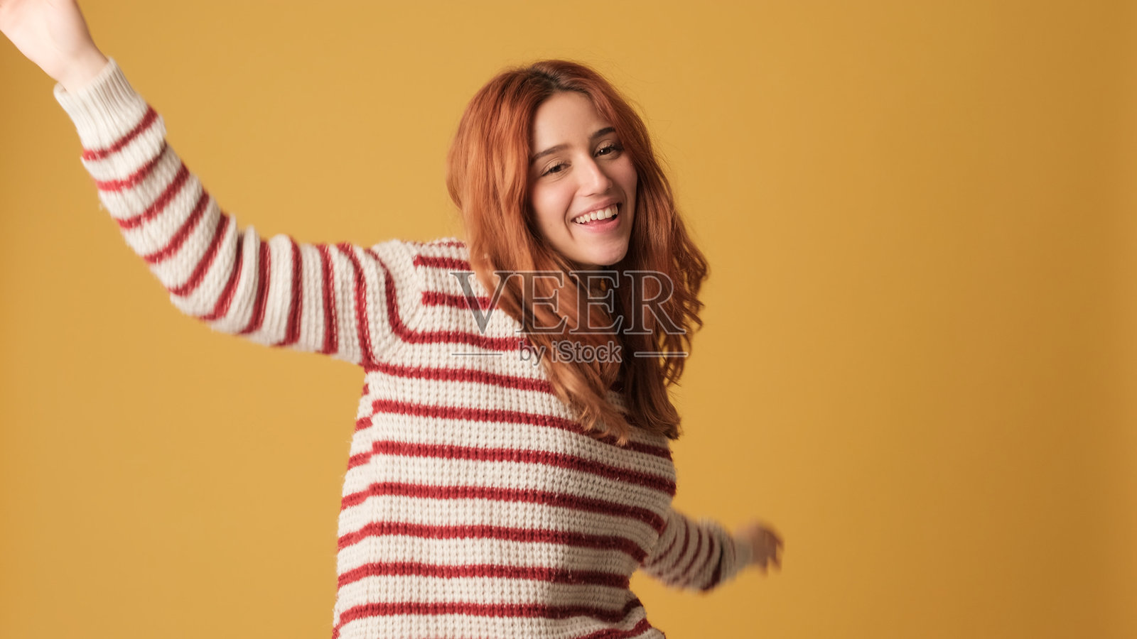 快乐的红发女性穿着毛衣，在黄色背景上尽情舞蹈。美丽的女孩在工作室中摆姿势，黄色墙面上留有空白空间供您进行宣传。照片摄影图片
