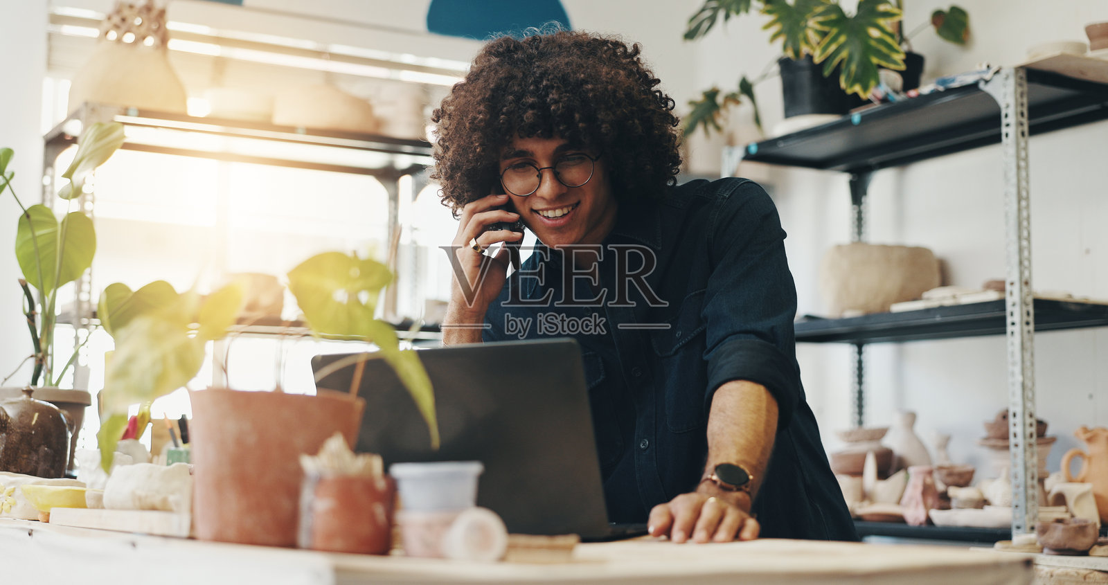 商人、笔记本电脑和陶艺店，接听客户服务、库存或在线订单的电话，位于陶土咖啡馆。男员工微笑着用手机与人交谈，适合小型创业企业。照片摄影图片