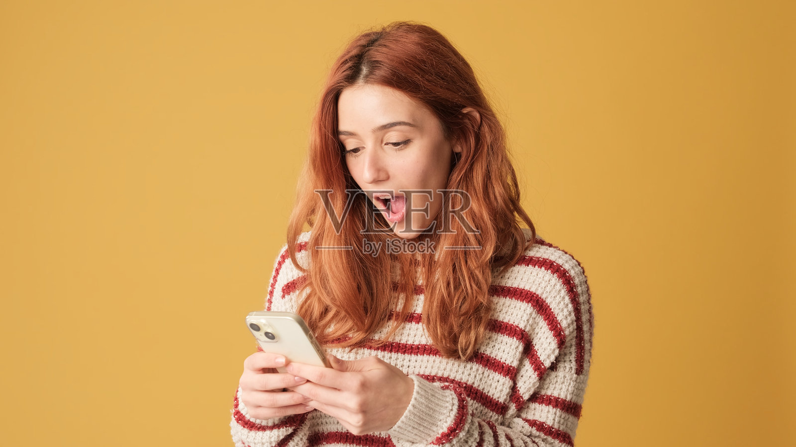 惊讶的红发女性穿着毛衣，手持智能手机对着镜头，背景为黄色。美丽的女孩在工作室中摆姿势，黄色墙面上有空白空间供您进行宣传。照片摄影图片