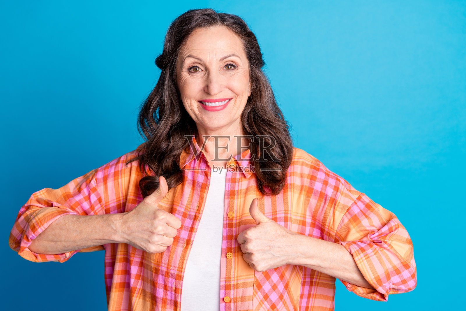 穿着格子衬衫的快乐成熟的女性在鲜艳的蓝色背景下竖起大拇指，展现出积极和快乐的情绪。照片摄影图片