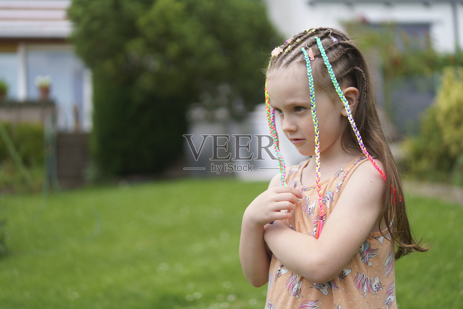 一个穿着裙子、扎着五颜六色小辫子、缺了一颗门牙的可怜小女孩，孤独而伤心，坐在夏天的后院，周围环绕着绿树和鲜花。这是健康的悲伤和情感的概念。照片摄影图片