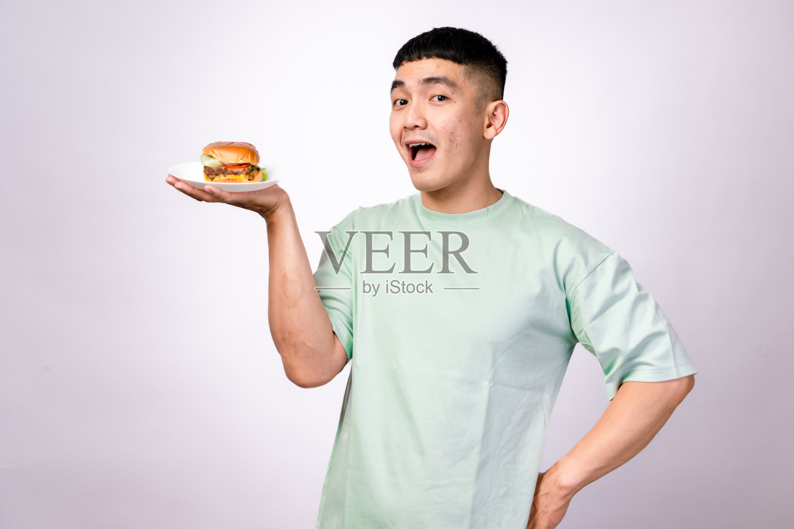 一位穿着薄荷绿衬衫的亚洲男子兴奋地在白色盘子上展示一个芝士汉堡。他面带灿烂的笑容，背景是纯白色的。照片摄影图片