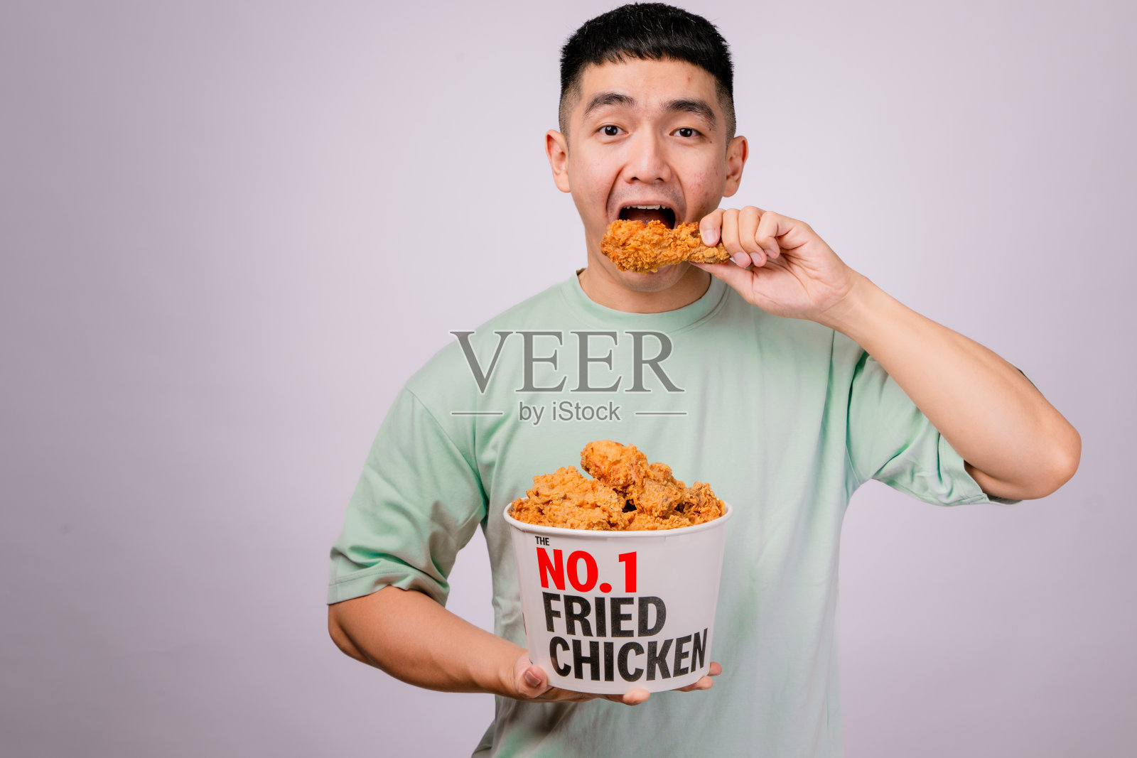 一位穿着浅绿色衬衫的亚洲男子正在享用一根酥脆的炸鸡腿，他手里拿着一个标有“第一炸鸡”的大桶。他看起来开心而满足。背景是白色的。照片摄影图片
