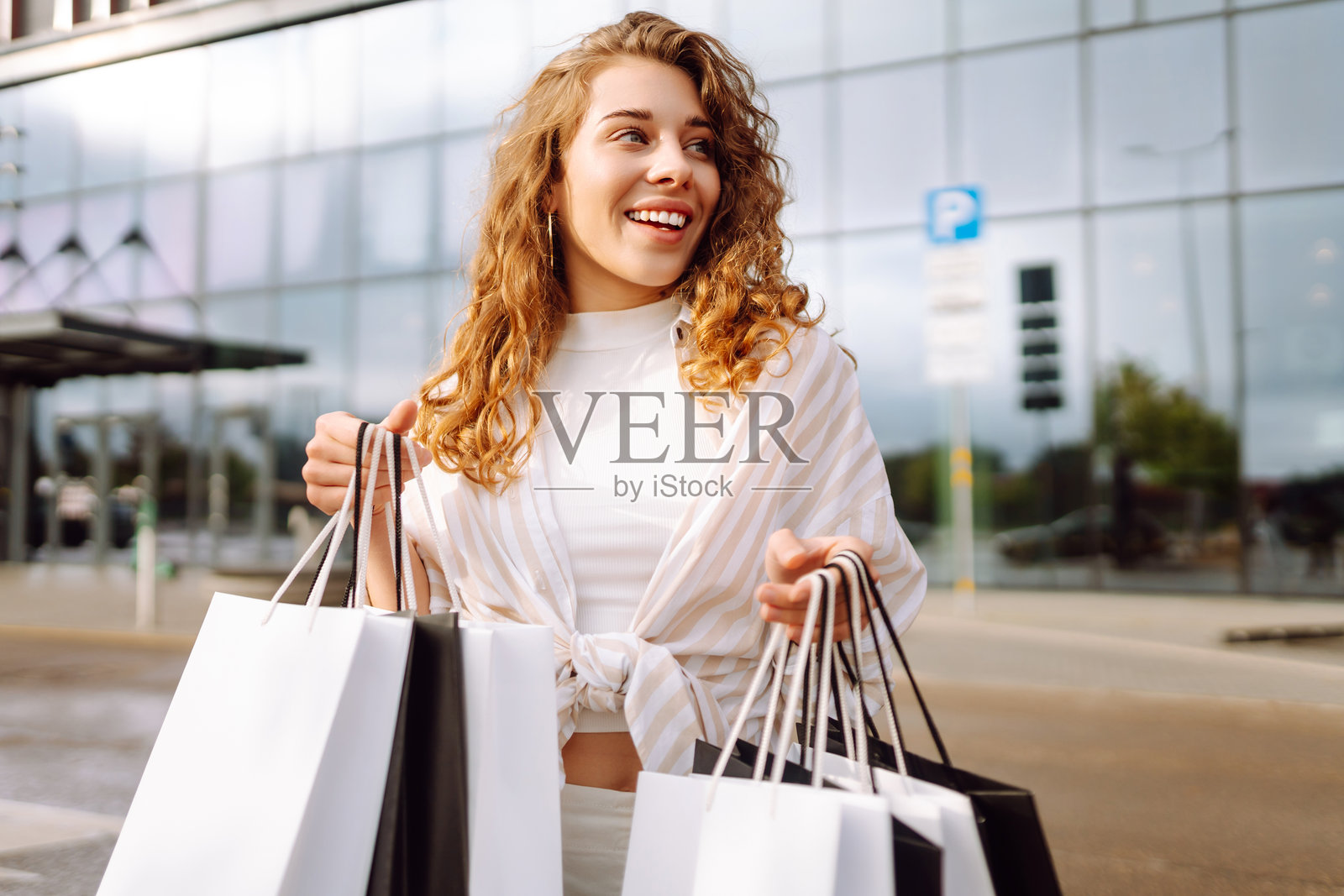 年轻女性穿着时尚衣服，卷发，手里提着购物袋，走在购物中心附近。照片摄影图片