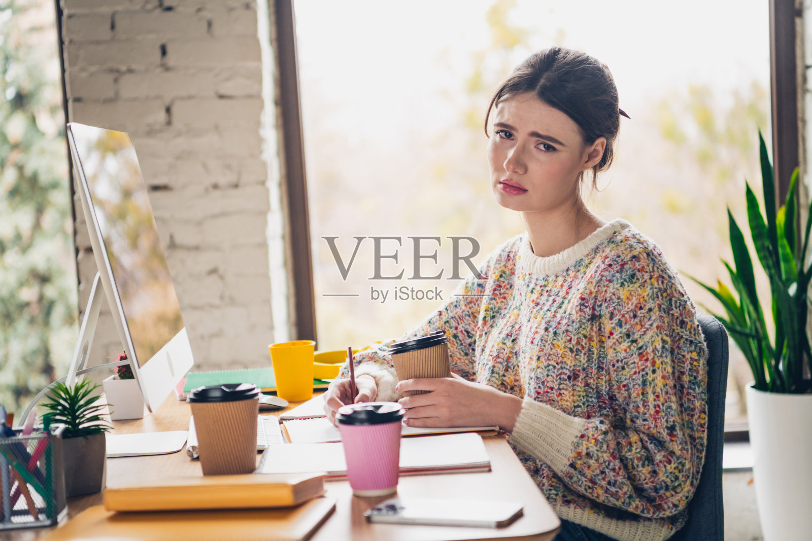 年轻女性坐在桌子旁，手握咖啡，面带沉思的表情，周围是五彩缤纷的物品，营造出温馨的家庭工作空间。照片摄影图片
