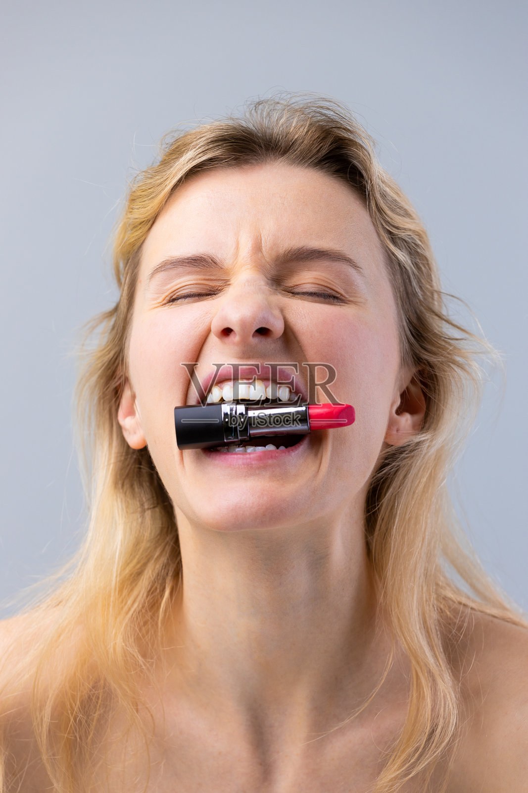 一个充满趣味和大胆的化妆冒险：一位女性兴奋地展示一支鲜艳的红色口红，嘴里叼着它，面带有趣的表情，背景则显得柔和。照片摄影图片