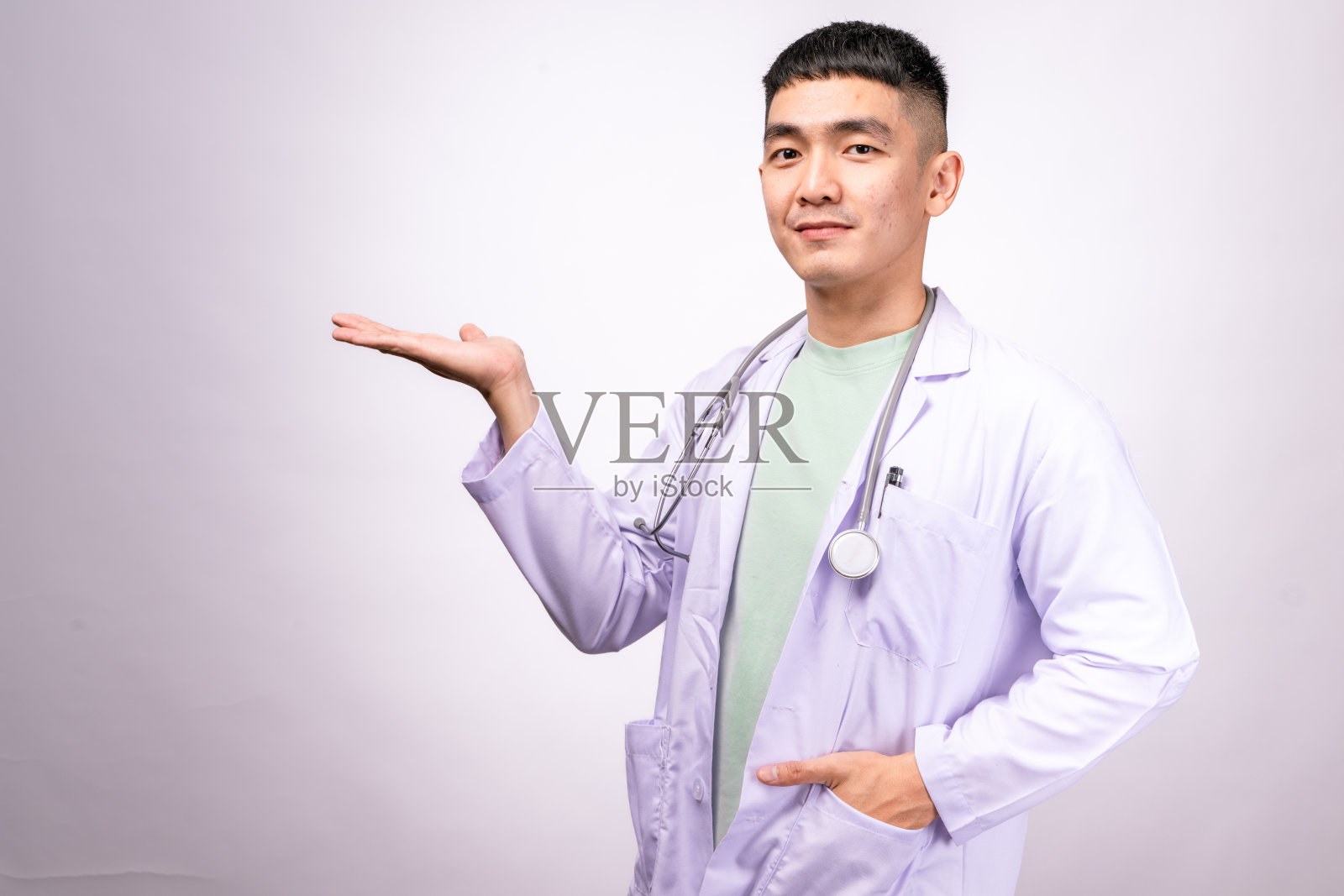 一位穿着白色实验室外套和听诊器的亚洲男性医生，手势如同在展示某物。他面带自信和友好的表情，背景为白色。照片摄影图片