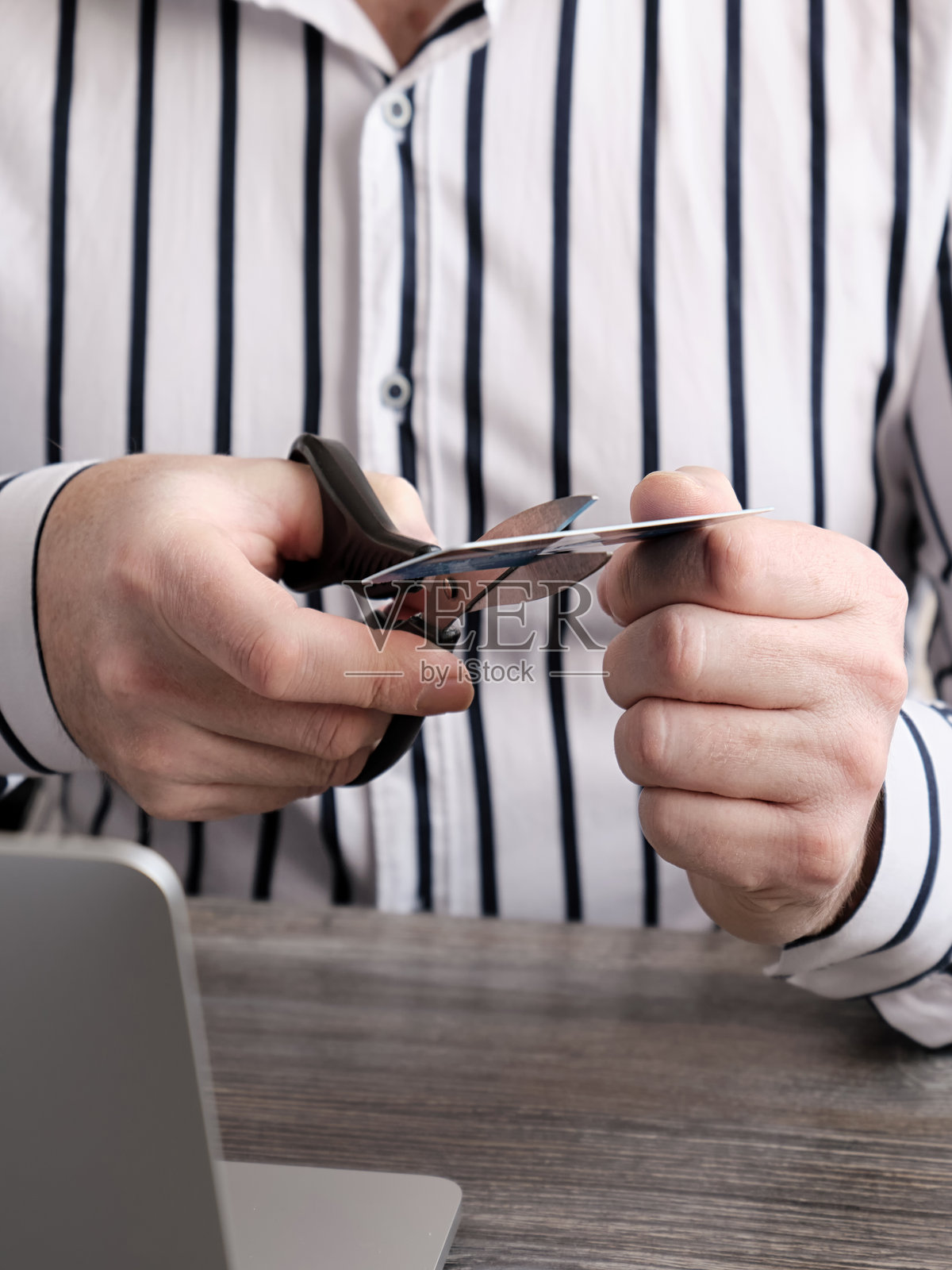 特写镜头：一只男人的手在笔记本电脑背景下用剪刀剪断一张塑料银行卡。照片摄影图片