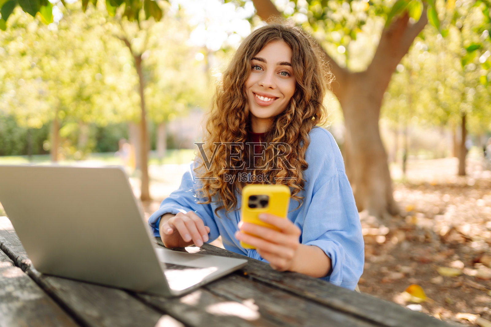 年轻女子坐在公园的木桌旁，手中拿着笔记本电脑和手机，卷发飘逸。照片摄影图片