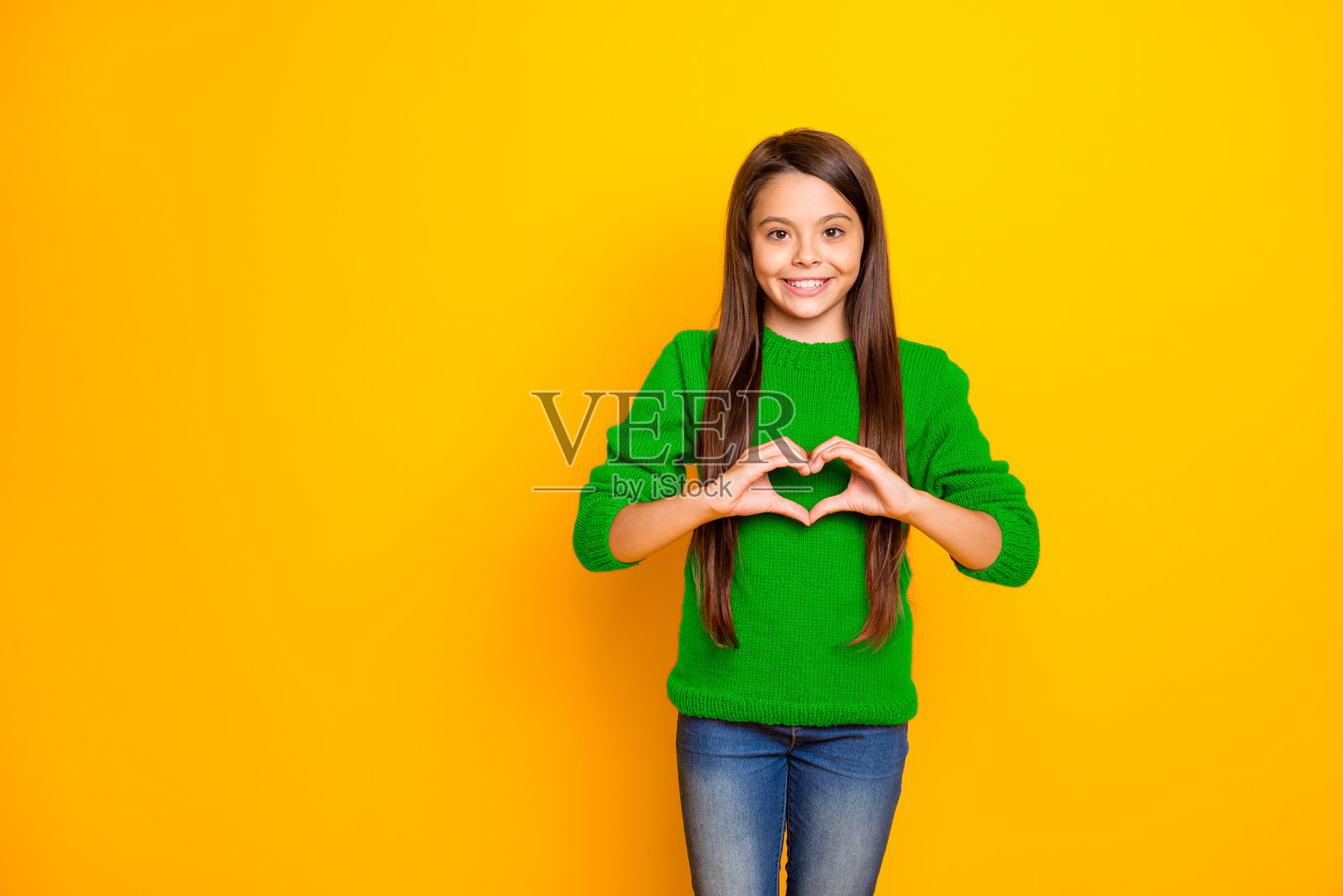 快乐迷人的女学生在明亮的黄色背景下，微笑着露出洁白的牙齿，穿着牛仔裤，做出心形手势。照片摄影图片