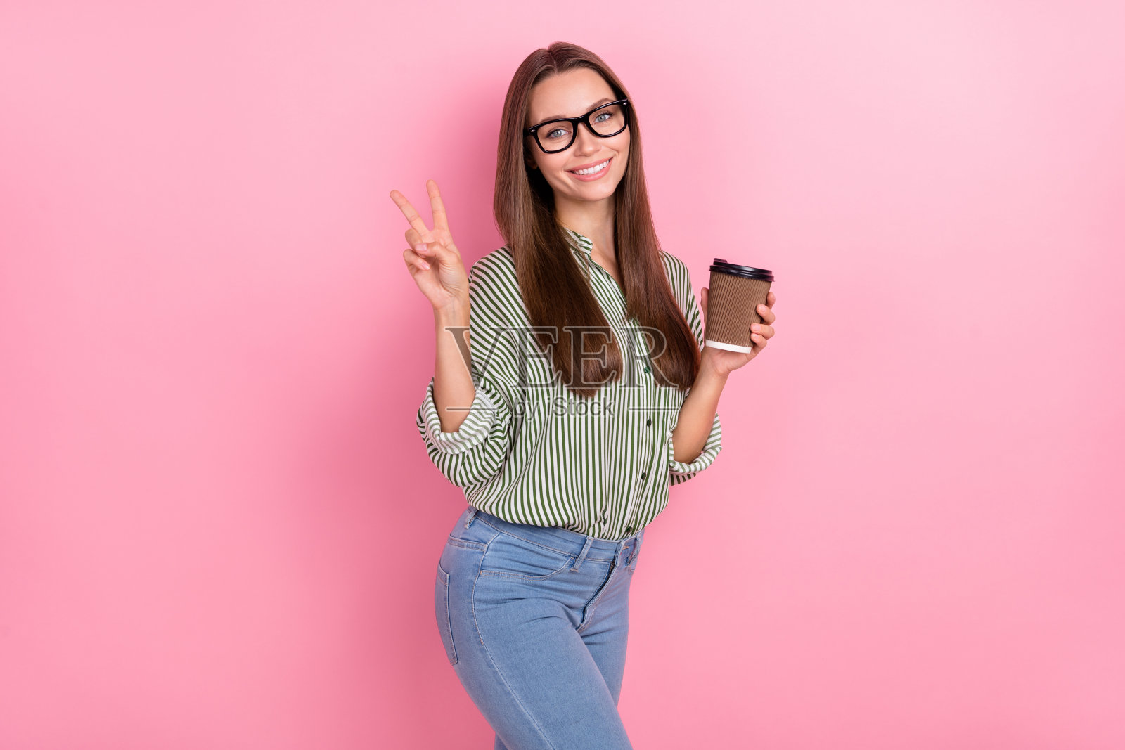 穿着绿色衬衫、戴着眼镜的迷人搞笑女士在粉色背景下喝茶并做出V字手势照片摄影图片