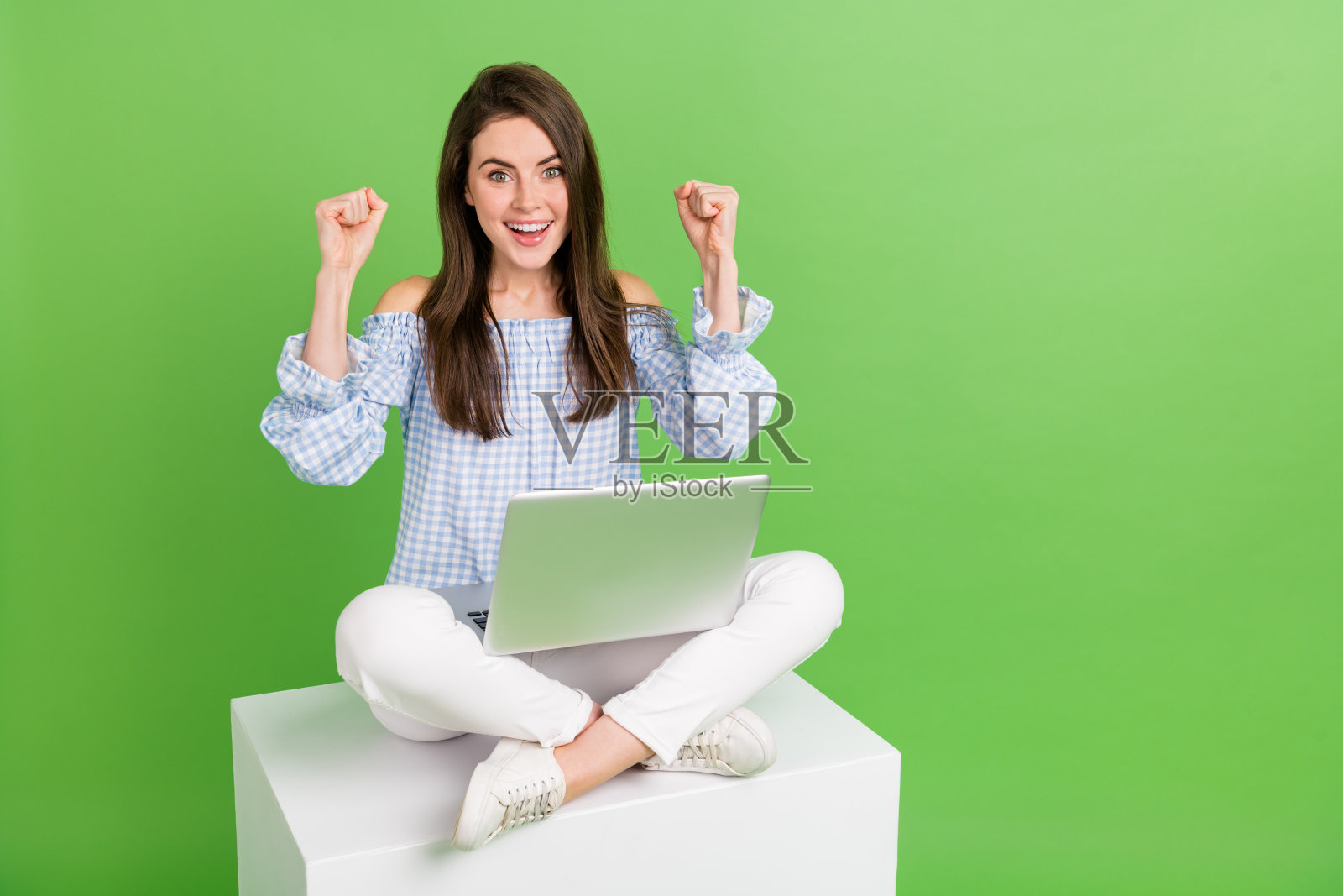 甜美心情的女孩坐在立方体平台上，举起拳头庆祝胜利，正在使用笔记本电脑，背景为绿色。照片摄影图片