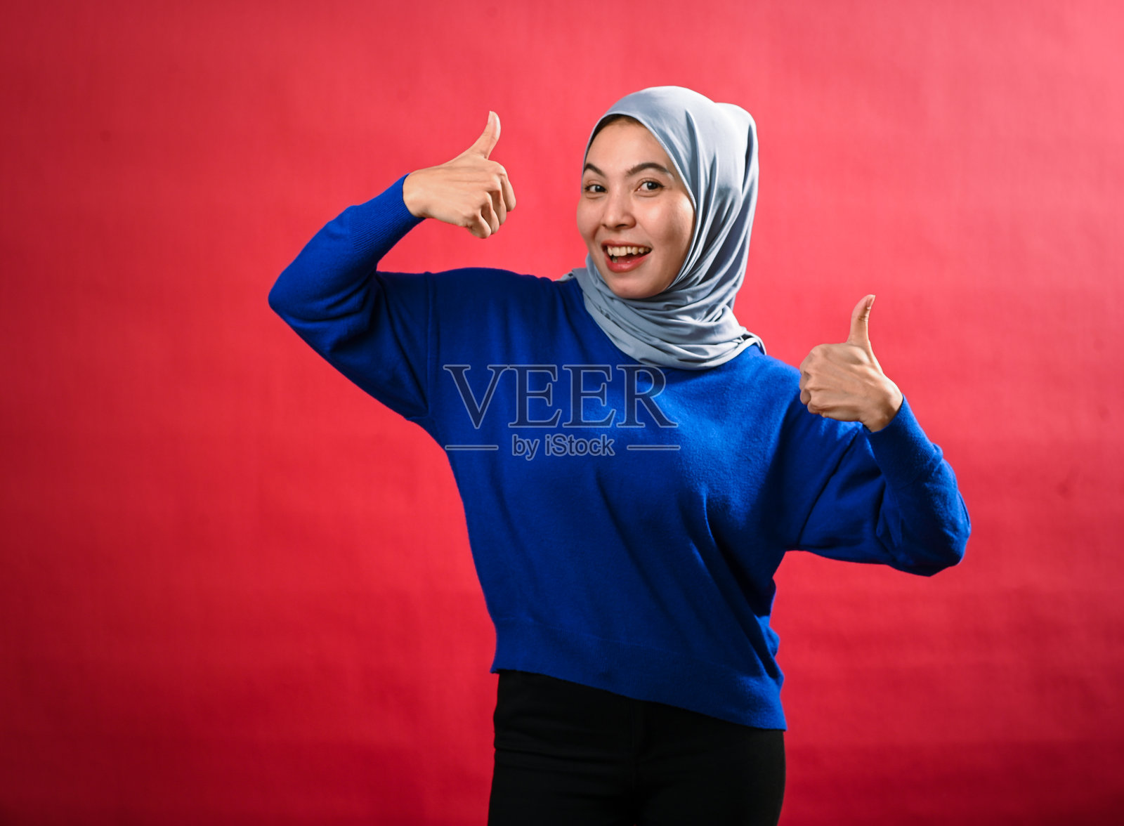一位亚洲女性穿着灰色头巾和蓝色毛衣，微笑着做出两个热情的竖起大拇指的手势。她自信地站在明亮的红色背景前，散发着积极的气息。照片摄影图片