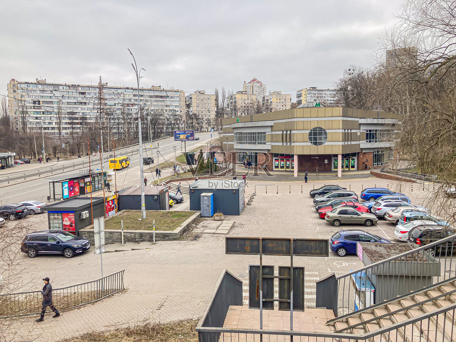 图像展示了一个城市街景，包含多层建筑、公交车站、铺砌区域、停放的车辆、散落的人群、阴云密布的天空和光秃的树木。地点 - 乌克兰基辅，2024年3月15日。照片摄影图片