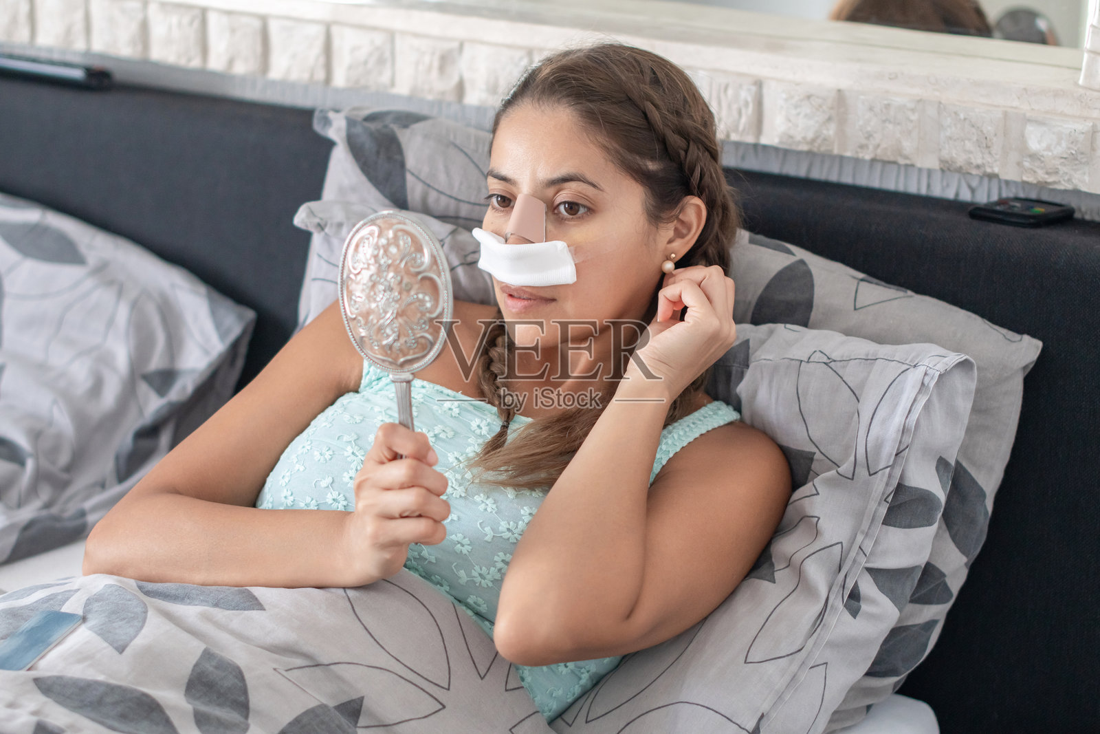 一位女性穿着家居服躺在床上，脸上贴着绷带，经过鼻子手术后，她看着镜子中的自己，整理头发，担心手术结果。照片摄影图片