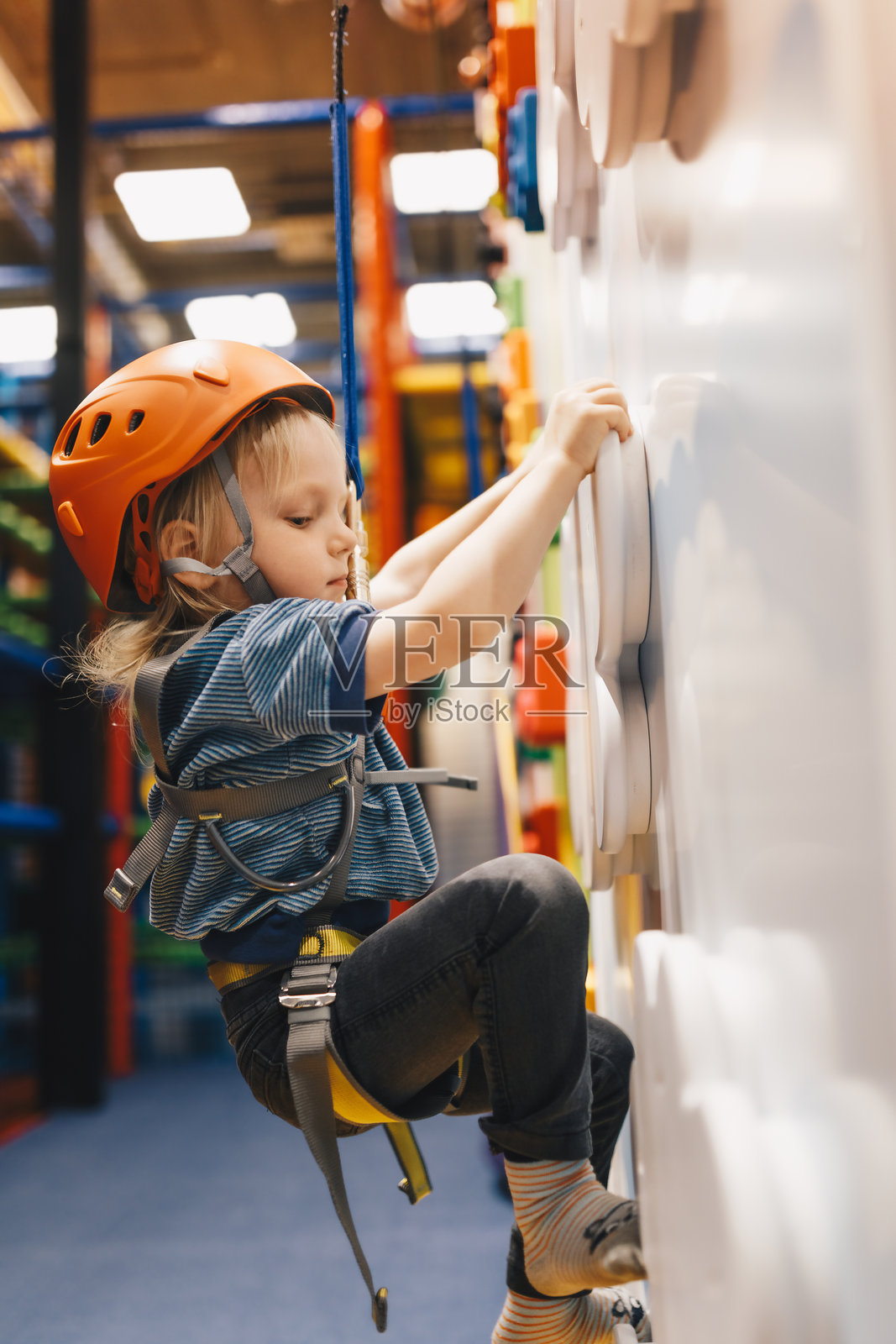 快乐的男孩戴着红色头盔在室内攀岩和抱石中心参加攀岩课程。男孩对着镜头微笑并竖起大拇指。照片摄影图片