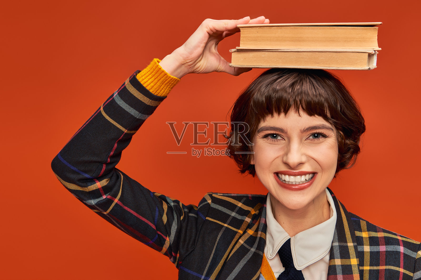 一位充满活力的年轻女性在橙色背景下，头顶平衡着书本，面带愉悦的微笑。照片摄影图片