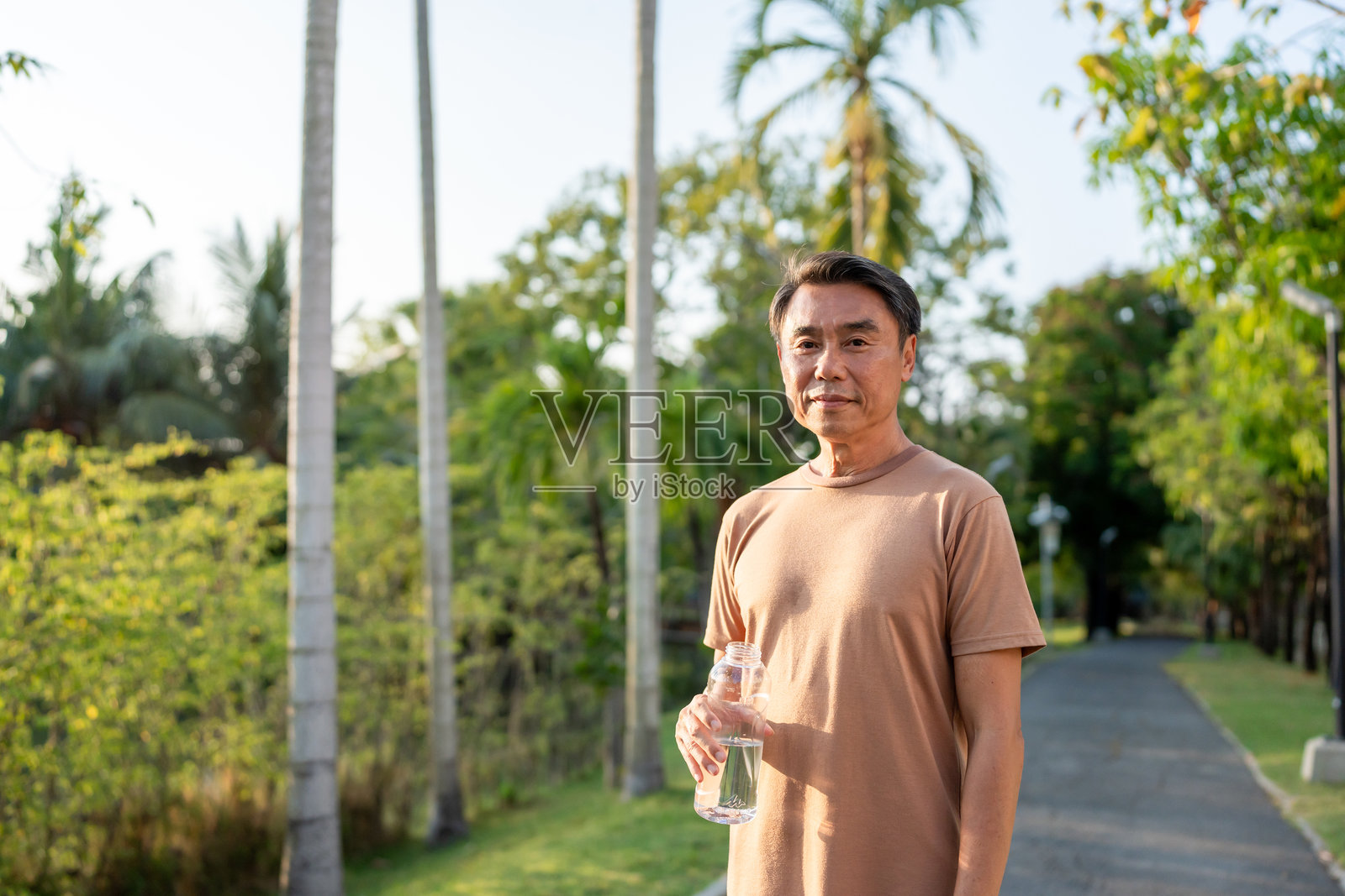 亚洲老年男子在花园里跑步时喝水的肖像。迷人的成熟老年男性慢跑者在日落时分的公共公园中锻炼后休息，注重健康。照片摄影图片