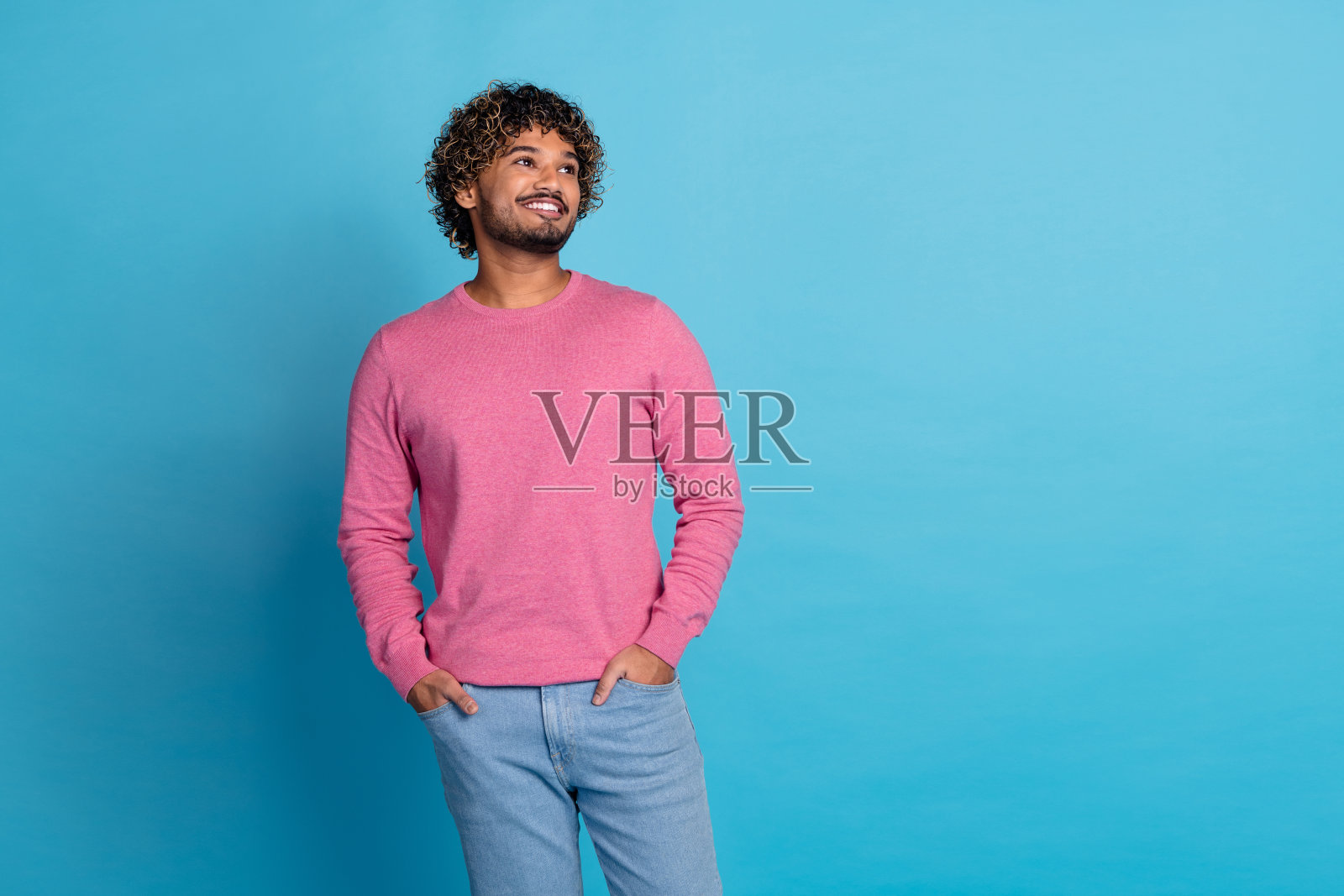 穿着休闲粉色毛衣的快乐年轻男子，卷发，自信地在鲜艳的蓝色背景前摆姿势照片摄影图片