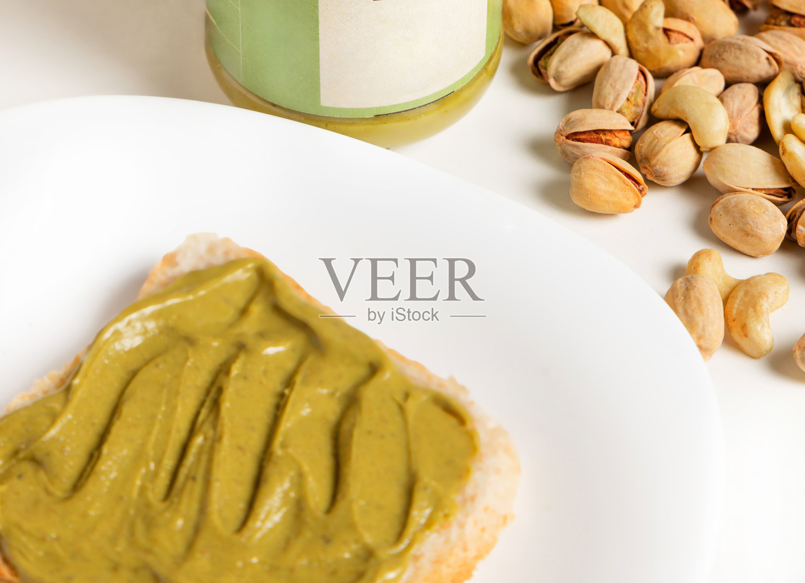 美味的开心果奶油涂抹在盘子或桌子上的吐司上。女人用厨房刀涂抹酱料，种子散落在白色表面上。留有文本空间，适合广告横幅。照片摄影图片