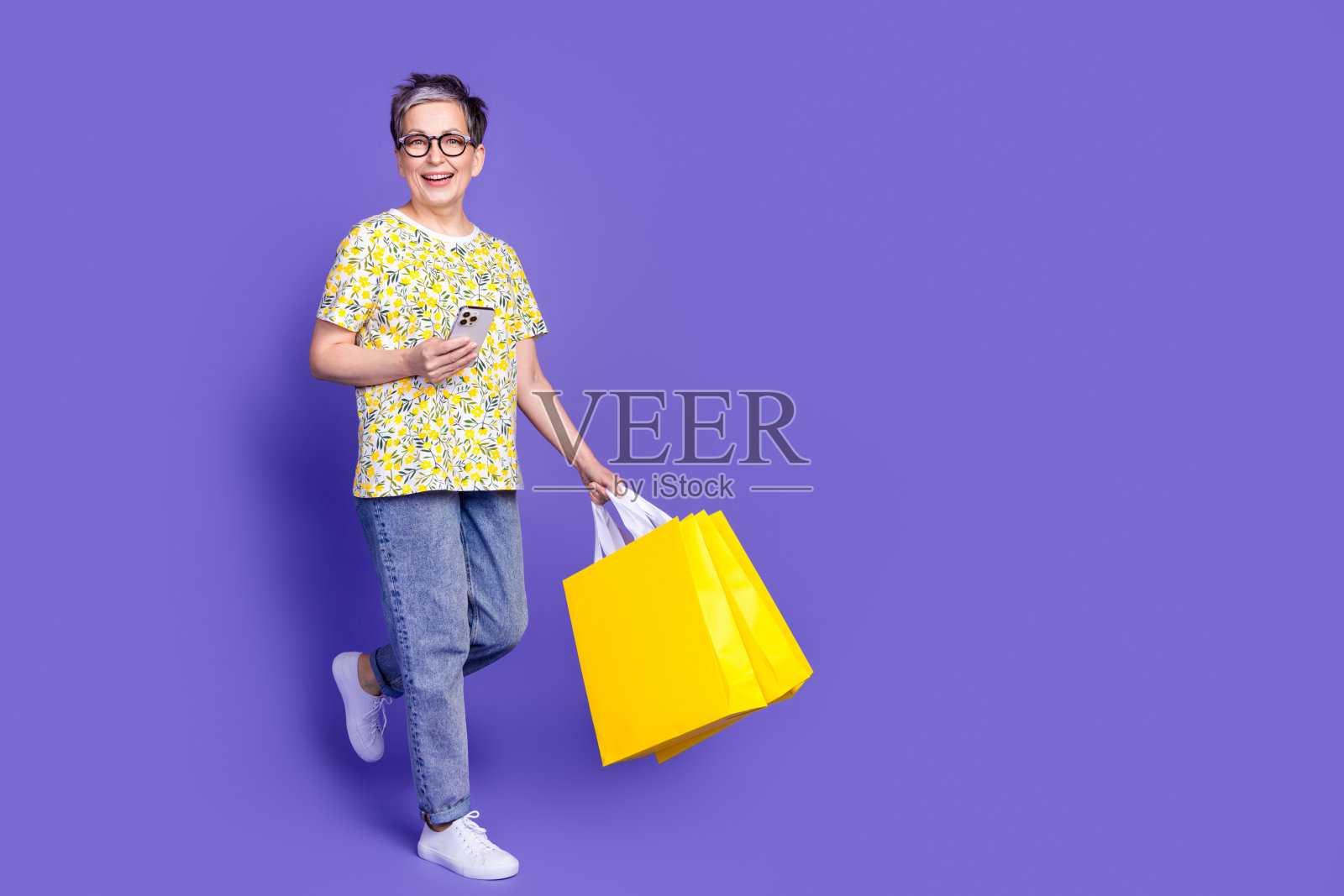 穿着时尚休闲服装的老年女性，手持购物袋和智能手机，背景为紫色。照片摄影图片