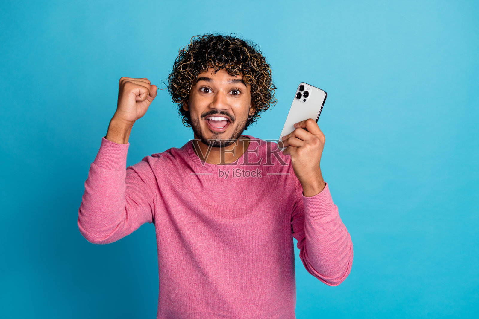 充满热情的年轻男子，卷发，手持智能手机，在鲜艳的蓝色背景下庆祝成功照片摄影图片