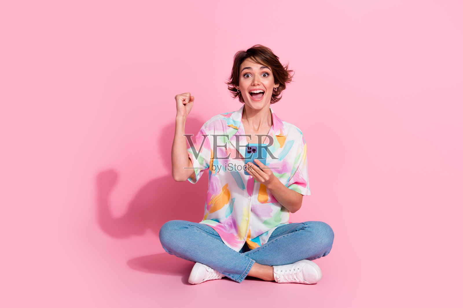 全身肖像，年轻女性举起拳头，手持智能手机，穿着衬衫，背景为粉色。照片摄影图片