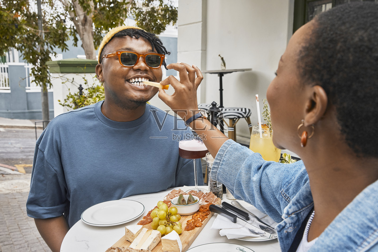 一位女性在社交聚会上给戴着太阳镜的男性朋友喂一片奶酪，享受着冷盘和开胃酒。黑人朋友们在食物和饮料中庆祝彼此的联系、欢笑和团聚，享受着充满正能量的周末。照片摄影图片