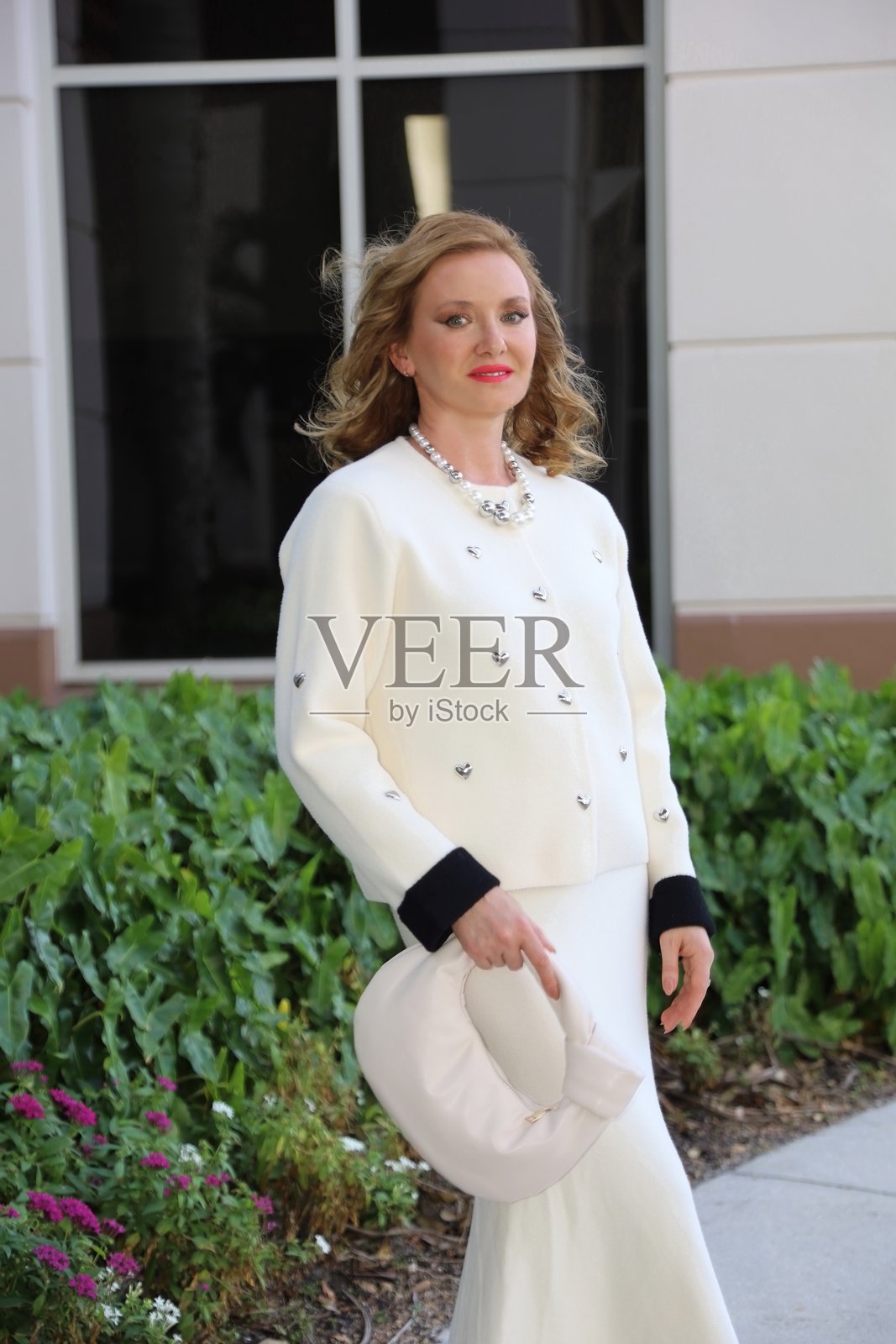 年轻美丽的女性穿着温暖的白色夹克和带黑色袖口的裙子，站在办公大楼前的铺砌道路上。照片摄影图片