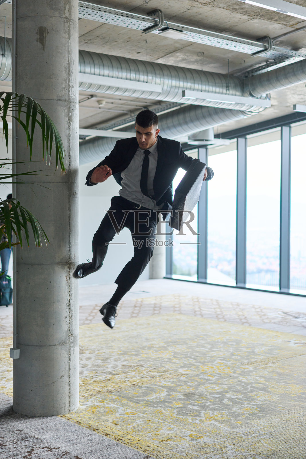 一位商人在现代办公室中兴奋地跳跃，手中拿着公文包，庆祝成功和胜利的时刻。照片摄影图片