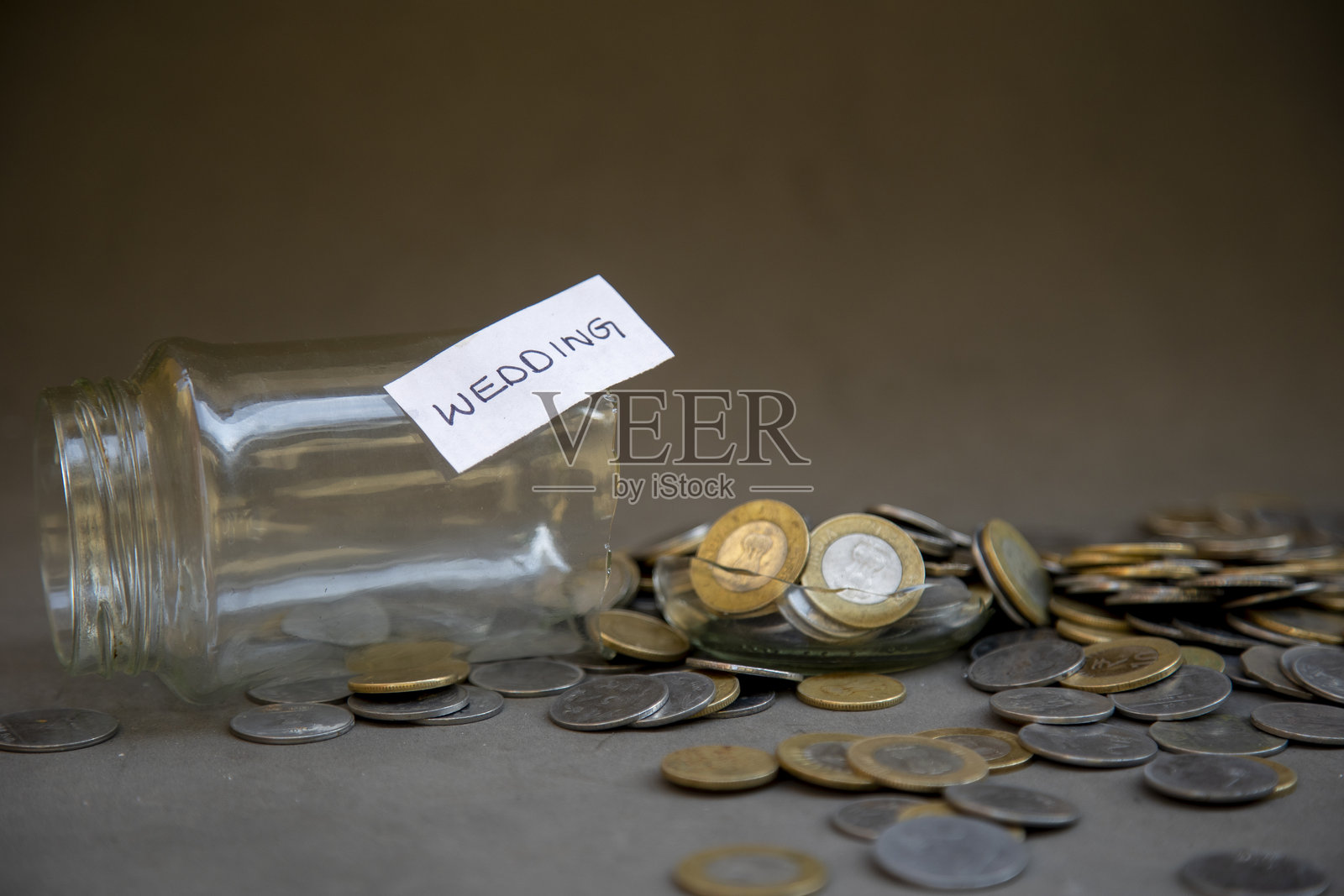 标有“婚礼”的玻璃罐破损，周围是象征经济压力的货币硬币照片摄影图片