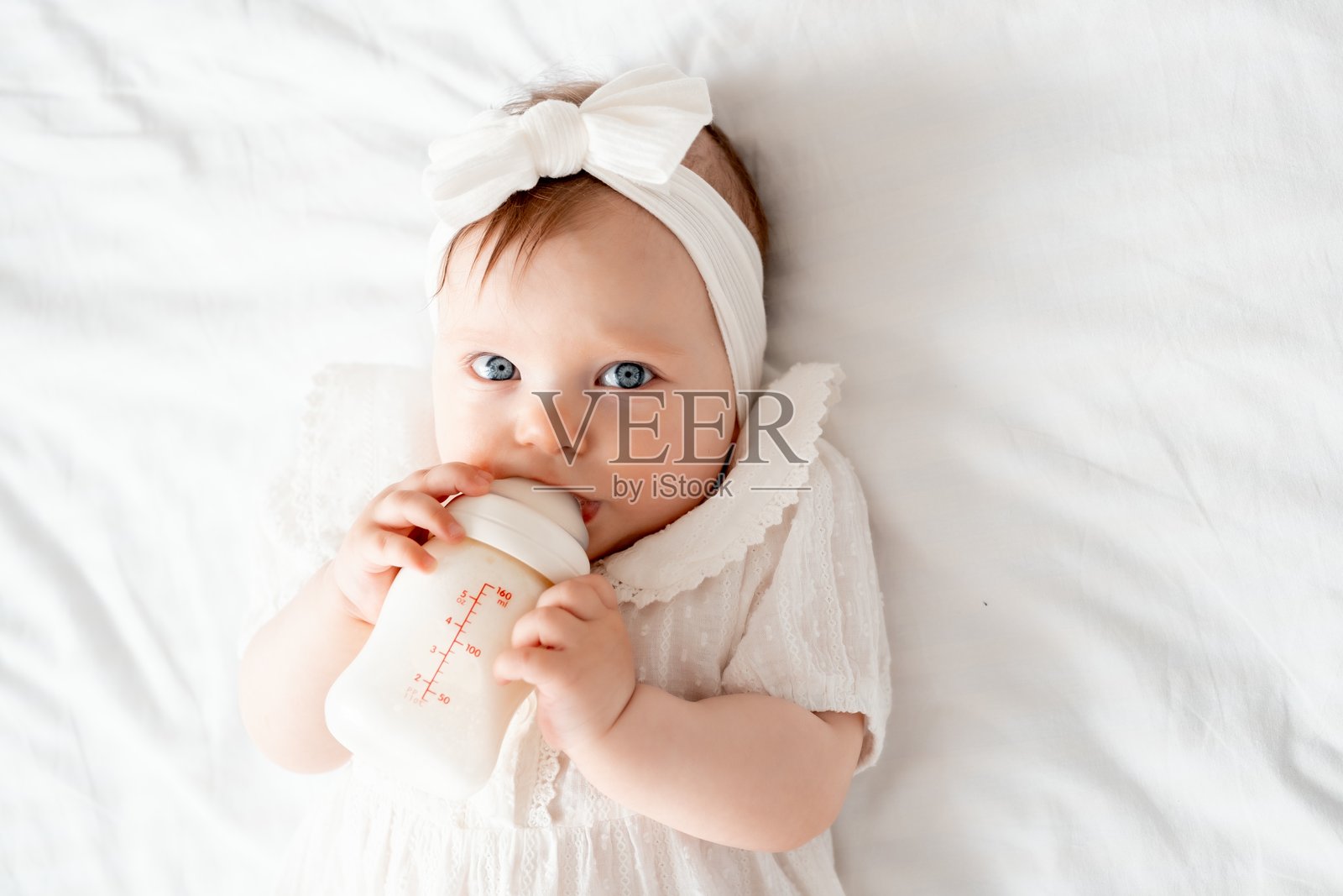 新生女婴穿着白色衣服仰卧在家里的床上喝奶瓶，可爱的宝宝在吃东西，留有文字空间照片摄影图片