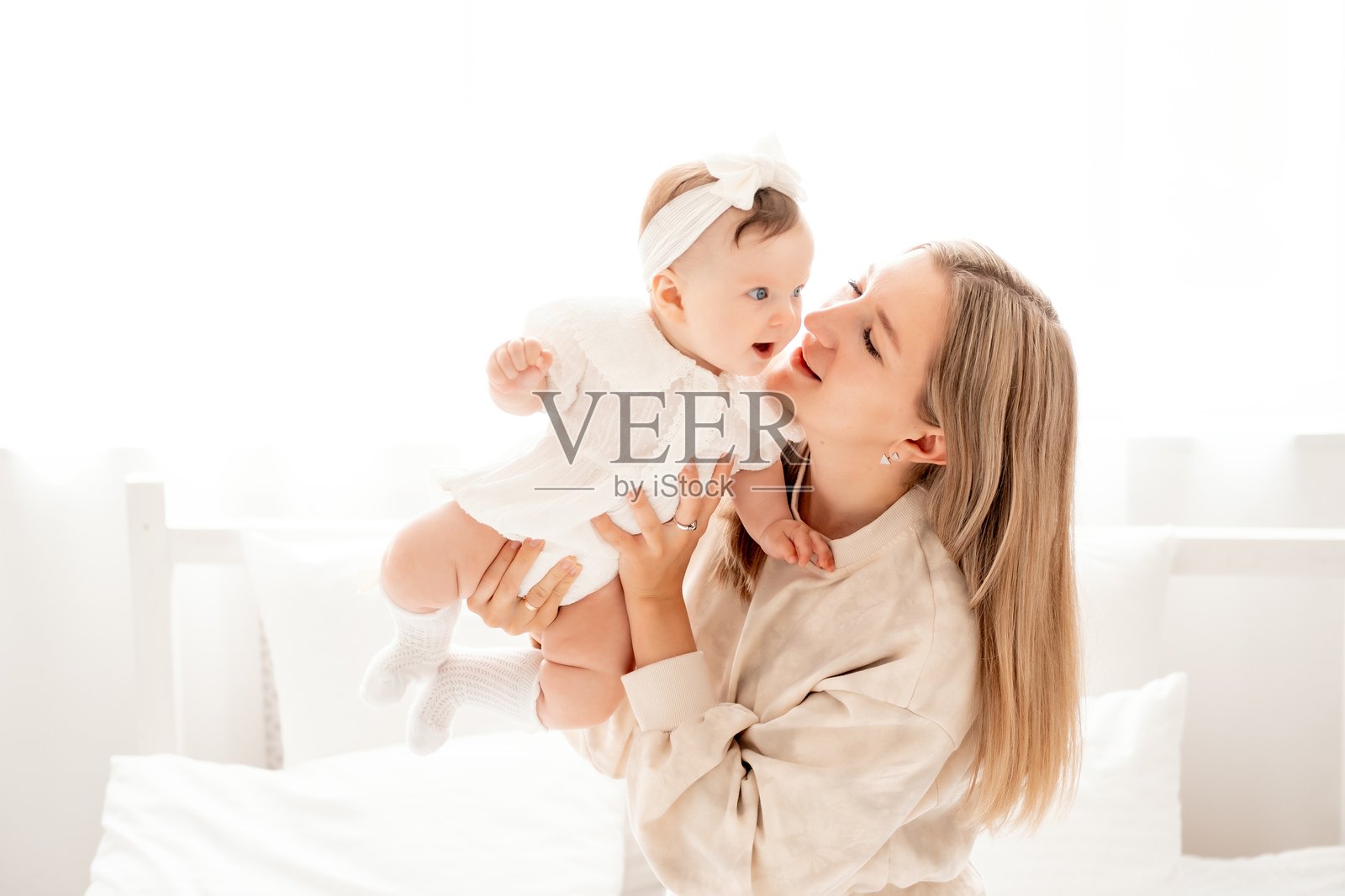 一位年轻母亲在家中白色床单或窗户前抱着新生儿，温柔地亲吻和拥抱她的宝宝，展现母爱，留有文字空间。照片摄影图片