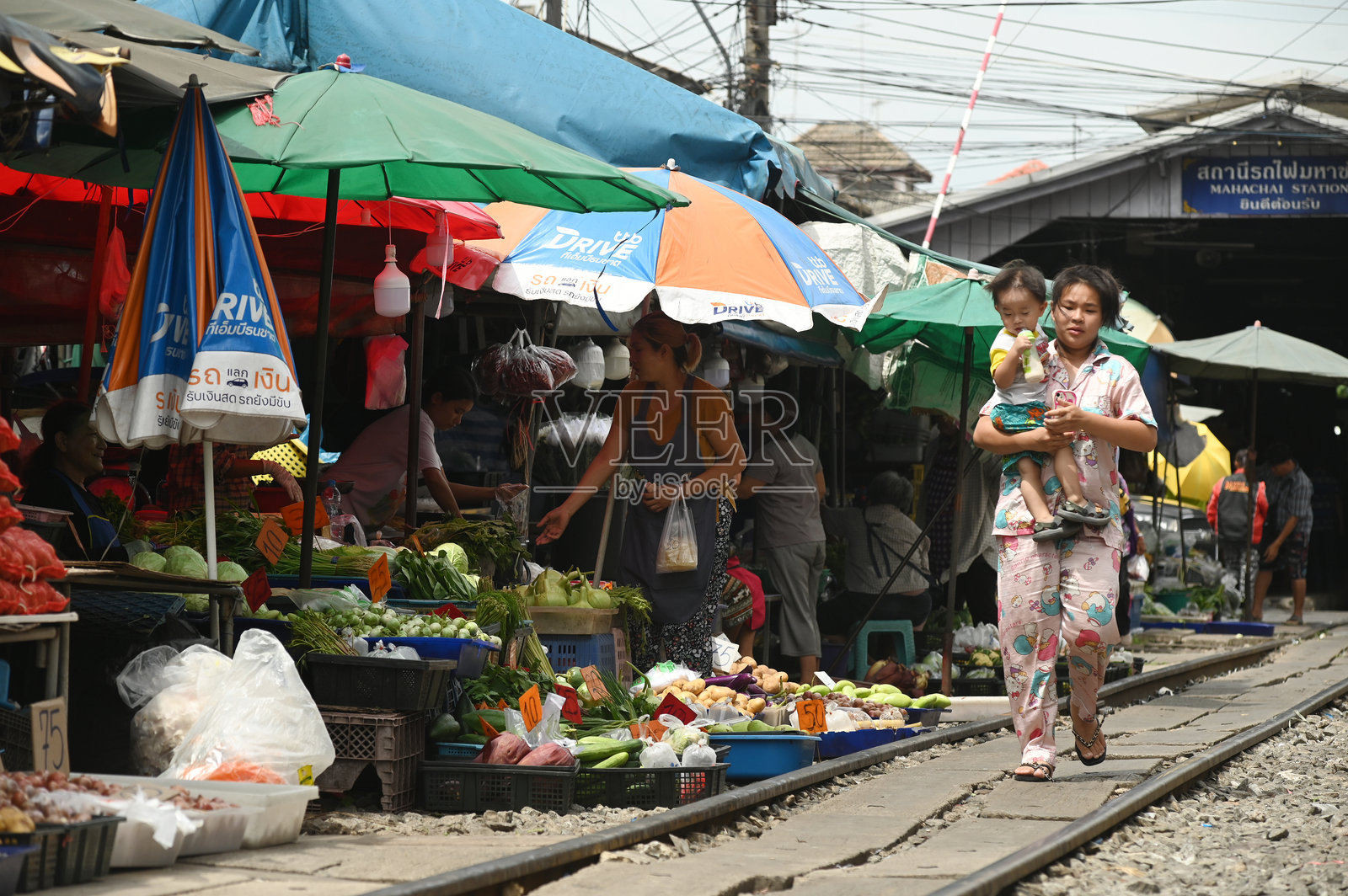 一位女性抱着她的孩子，沿着铁路轨道走，旁边是马哈猜车站的一个水果和蔬菜商店。照片摄影图片