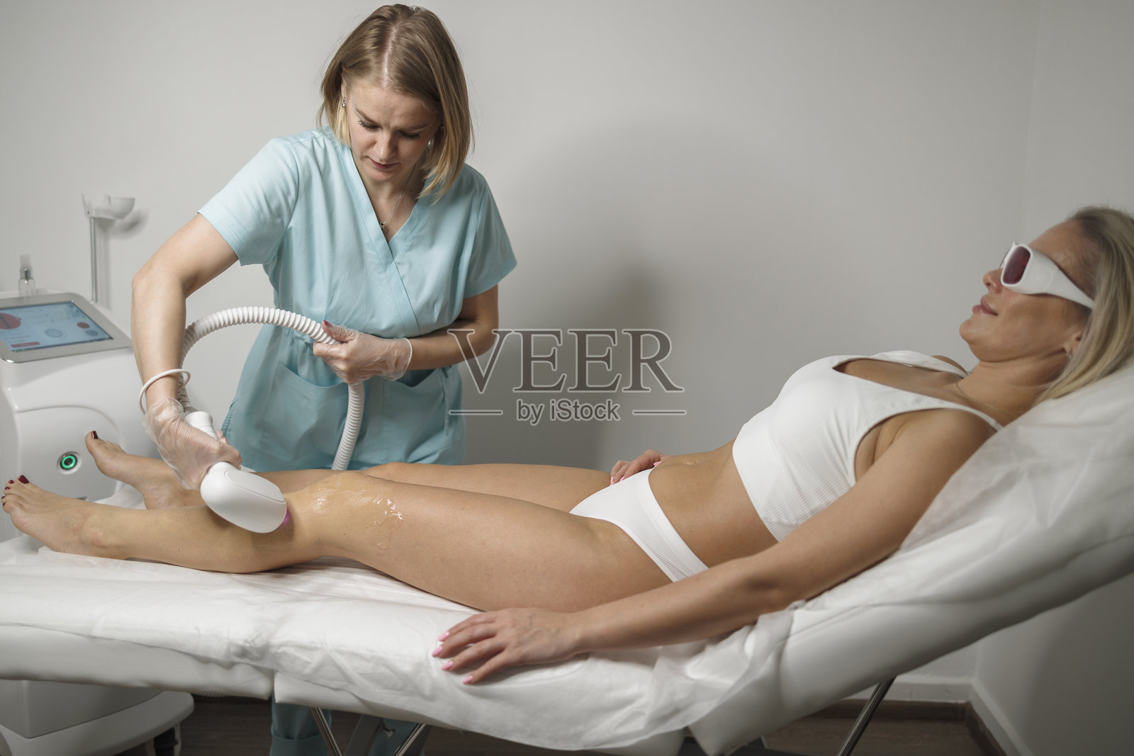 女性美容师正在为躺在水疗沙龙休闲椅上的金发女性进行激光脱毛治疗。照片摄影图片