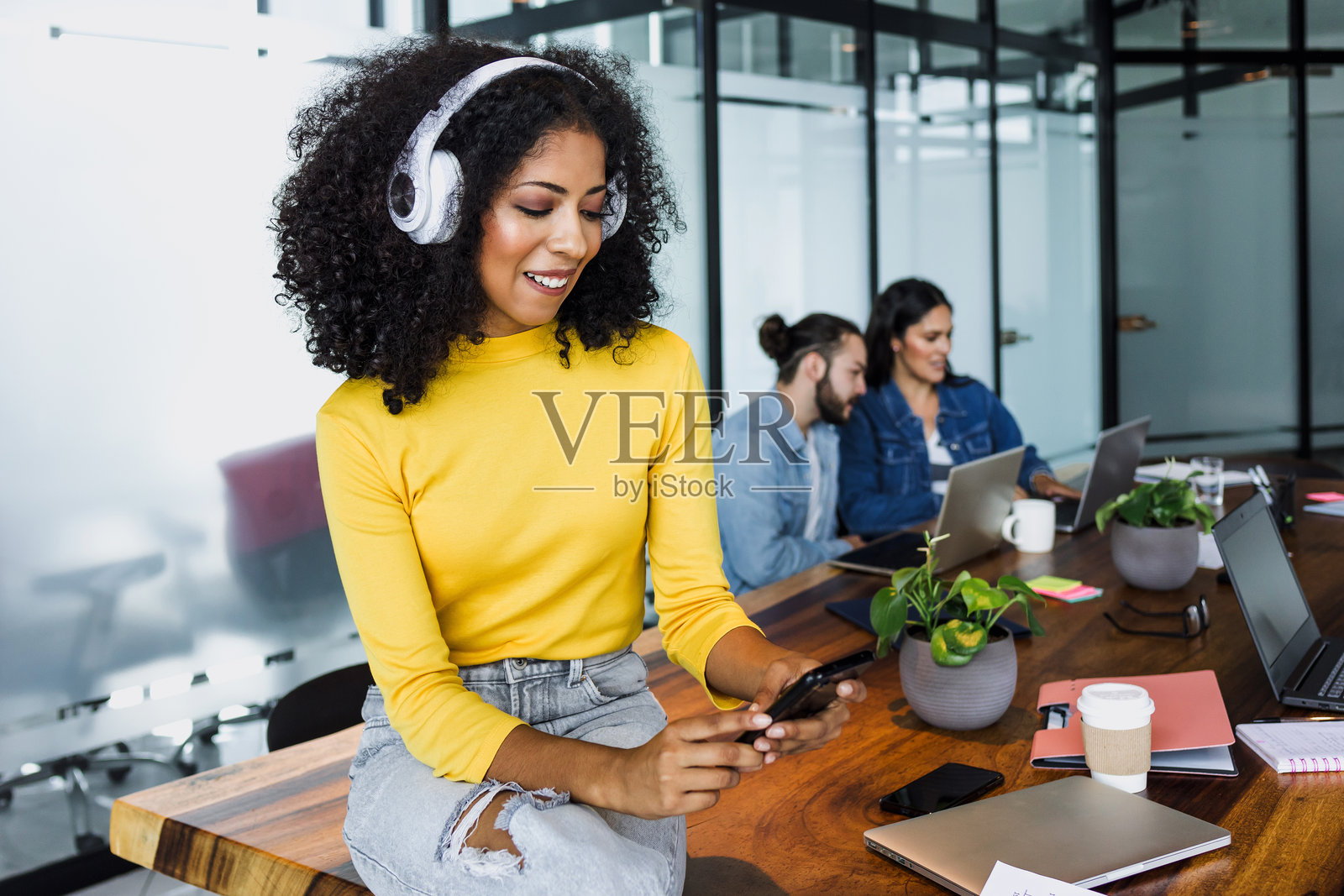 年轻的拉丁裔女性，留着非洲卷发或卷曲发型，戴着耳机在墨西哥的办公室里听音乐，手里拿着手机，非裔美国女性在工作场所。照片摄影图片