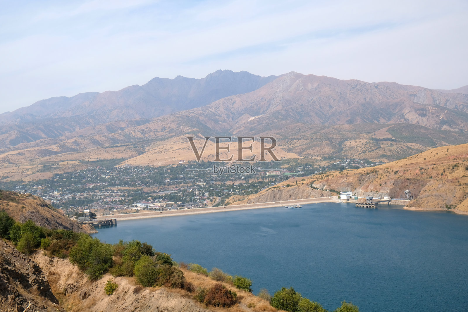 白天晴朗的蓝天下，宁静的湖泊和城市在山脉景观中尽收眼底。照片摄影图片