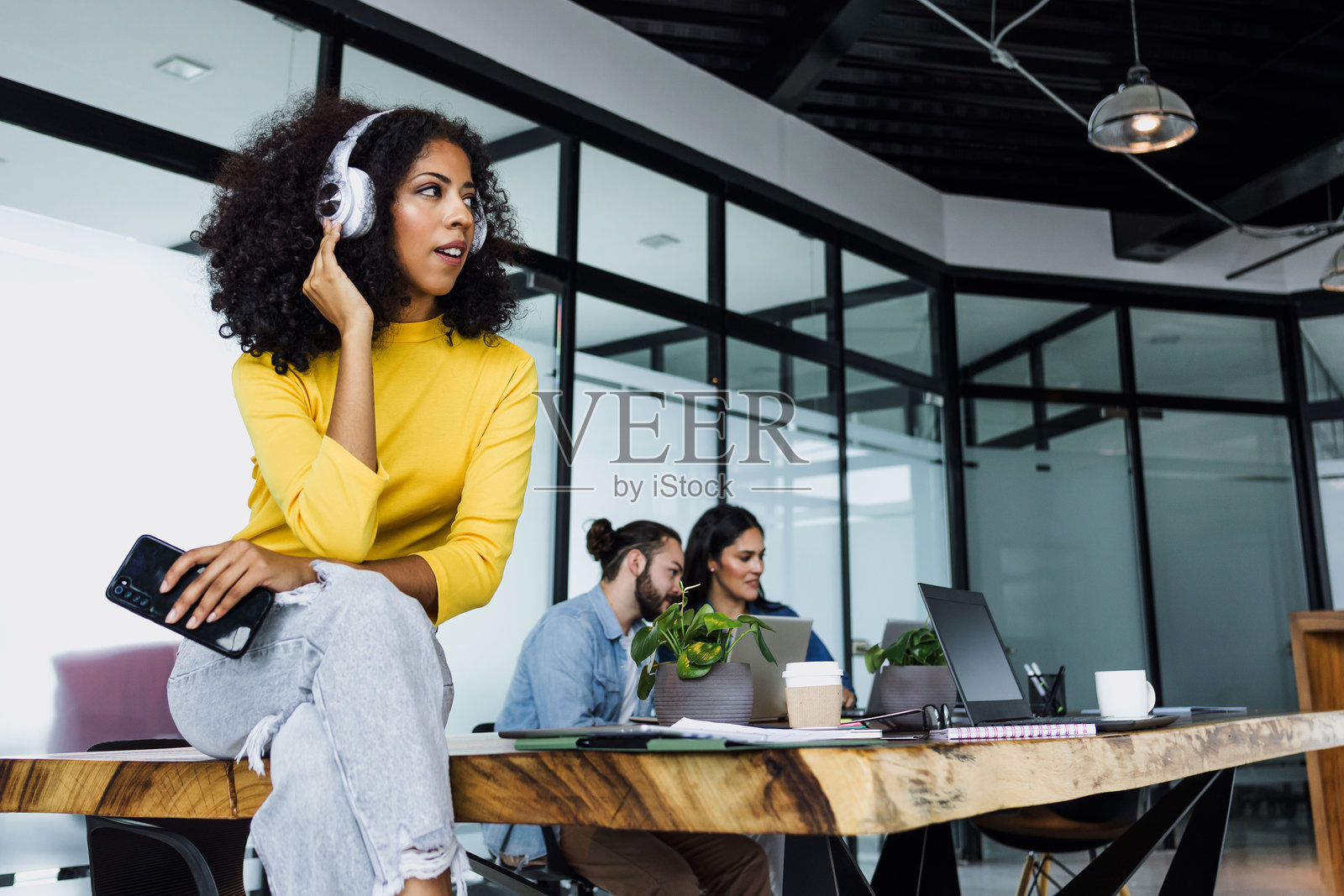 年轻的拉丁裔女性，留着非洲卷发或卷曲发型，戴着耳机在墨西哥的办公室里听音乐，手里拿着手机，非裔美国女性在工作场所。照片摄影图片