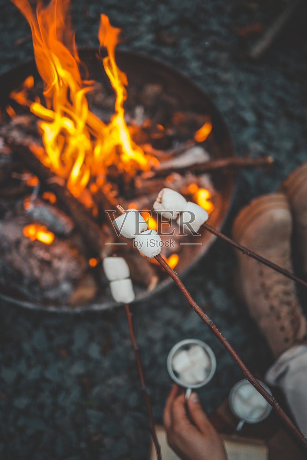 一群人在篝火上烤棉花糖，手里拿着棉花糖的棍子靠近明亮的火焰。热饮中也有棉花糖。他们享受着温馨的户外时光。照片摄影图片