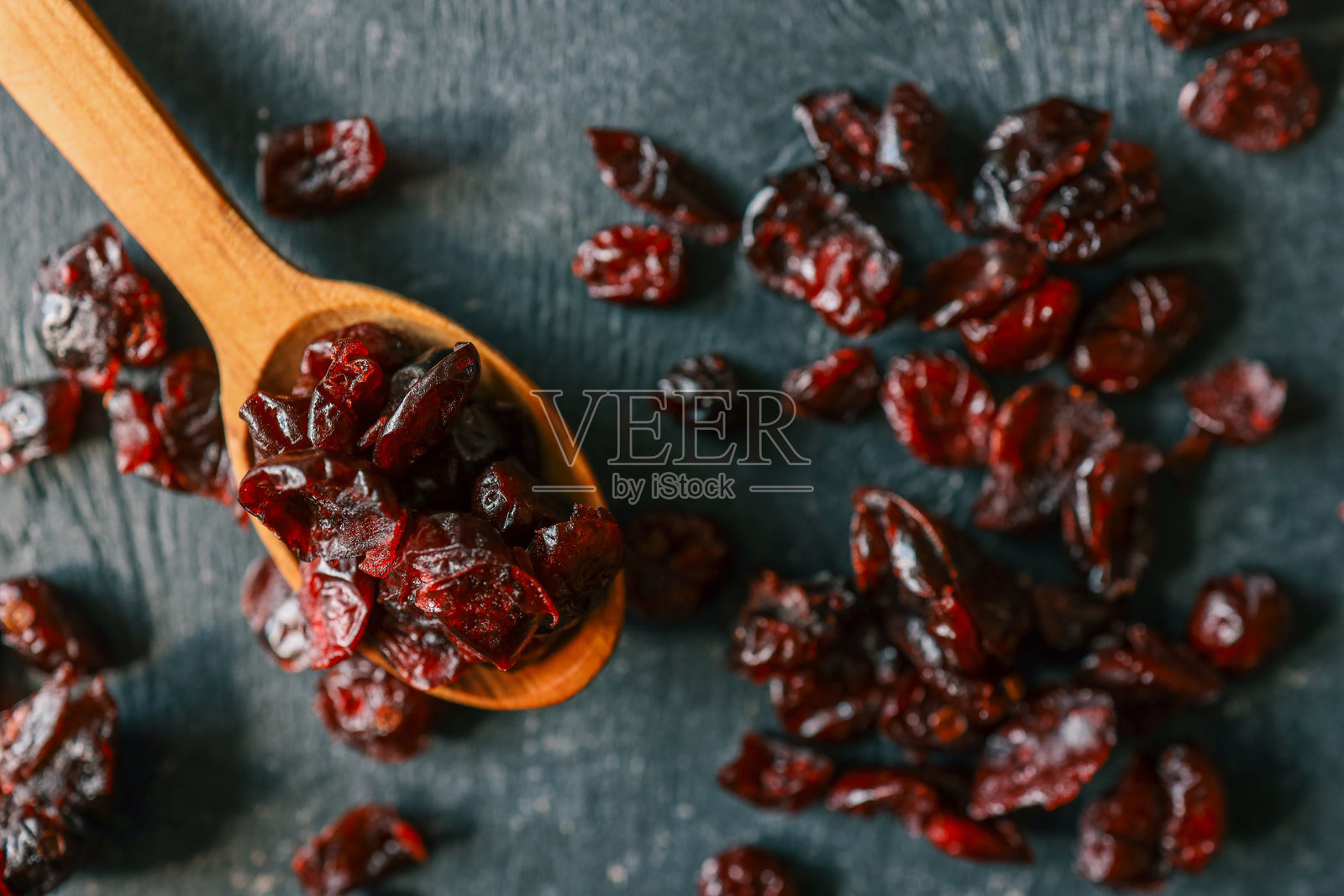 从上方俯视干蔓越莓，放在深色纹理表面上，旁边有一把木勺。光泽的红色浆果增添了丰富的色彩，常用于健康零食和烘焙食谱中。照片摄影图片