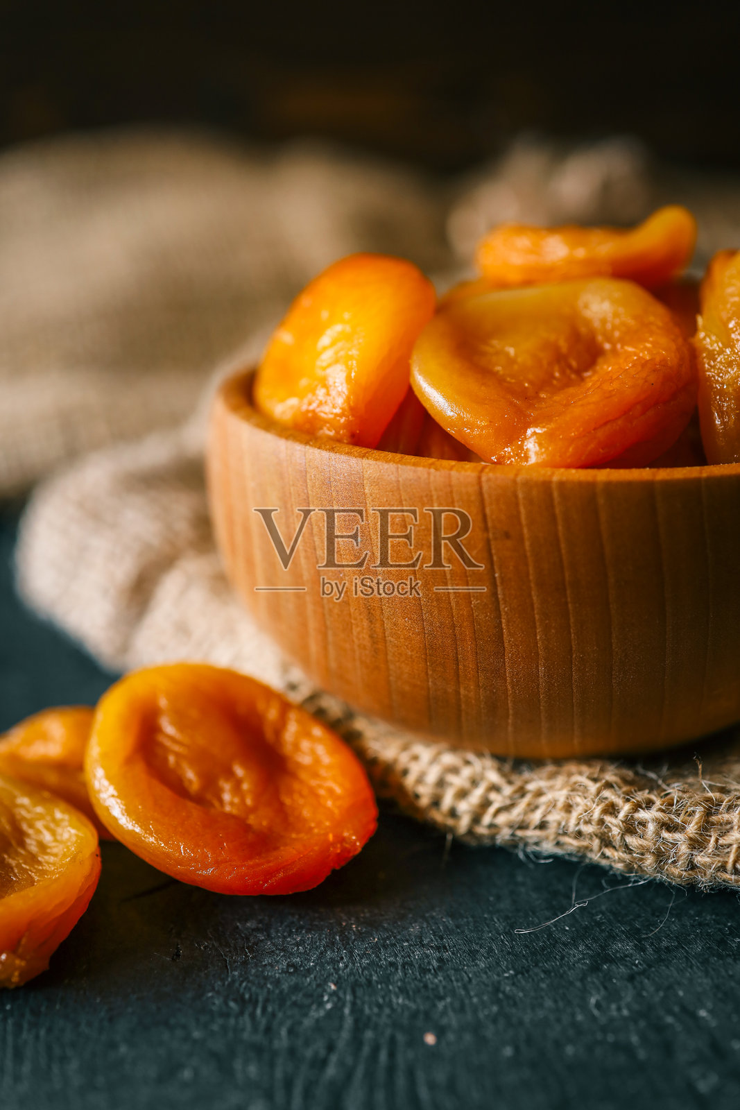 木碗和勺子里的干杏干，放在粗糙的麻布上。富含维生素和纤维的有机健康零食，在深色纹理背景下的自然温暖光线中拍摄。照片摄影图片