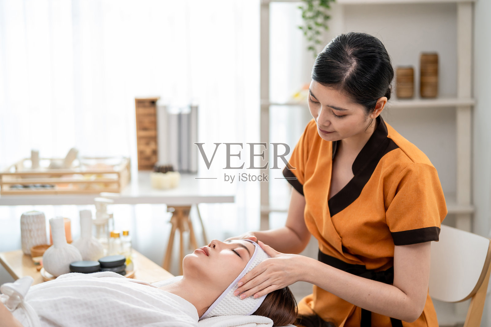 亚洲年轻女性在美容水疗中心接受头部和面部按摩治疗。迷人的女孩躺在按摩床上，享受按摩师提供的放松理疗服务，以护理身体。照片摄影图片