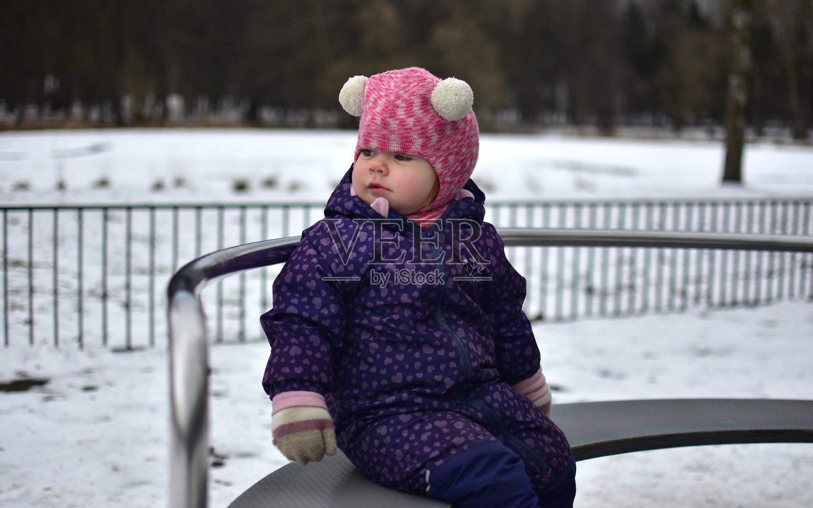 小女孩高兴地坐在游乐设施上，凝视着城市公园的儿童游乐场，秋天的寒冷天气中。幼儿女孩在冬季的公共游乐场玩得很开心。照片摄影图片