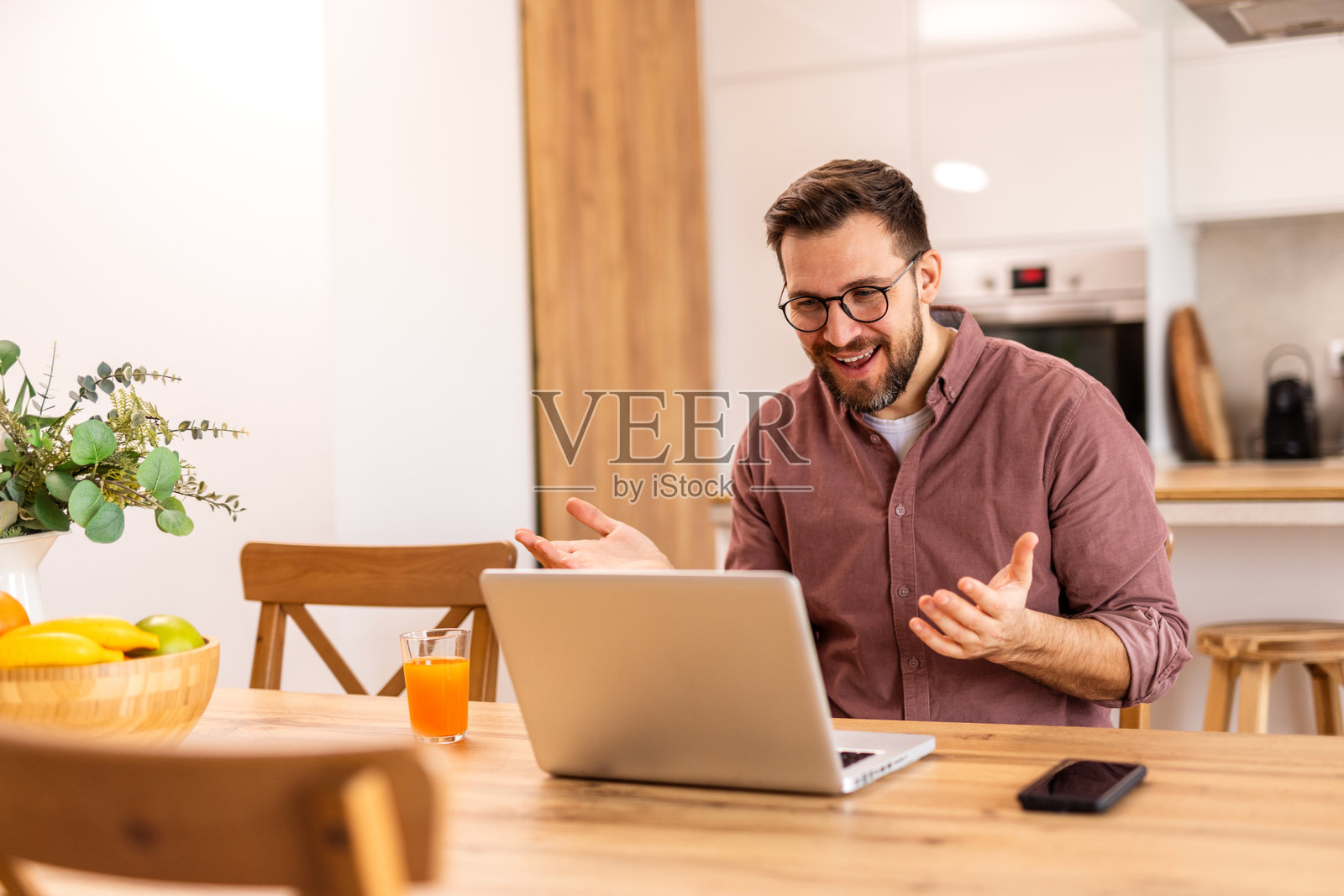 微笑的男子在家中用笔记本电脑工作，坐在木桌旁。温馨的客厅里有柔和的灯光、书架和一碗新鲜水果，营造出轻松而富有成效的氛围。照片摄影图片