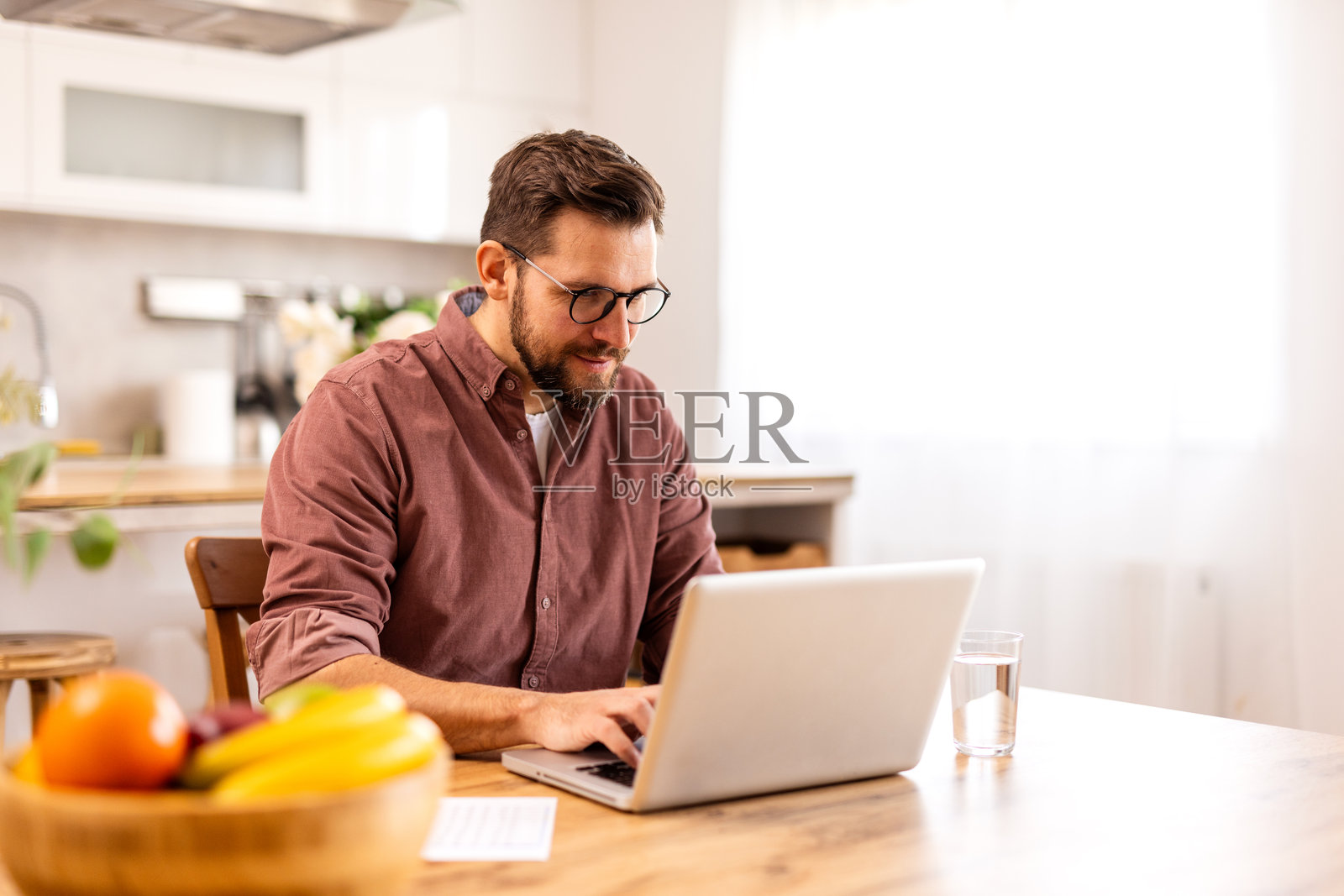 快乐的男人在舒适的家庭办公室里用笔记本电脑远程工作。温暖的灯光、书架和室内植物营造出一个舒适的工作空间。一碗水果增添了健康生活的气息。照片摄影图片