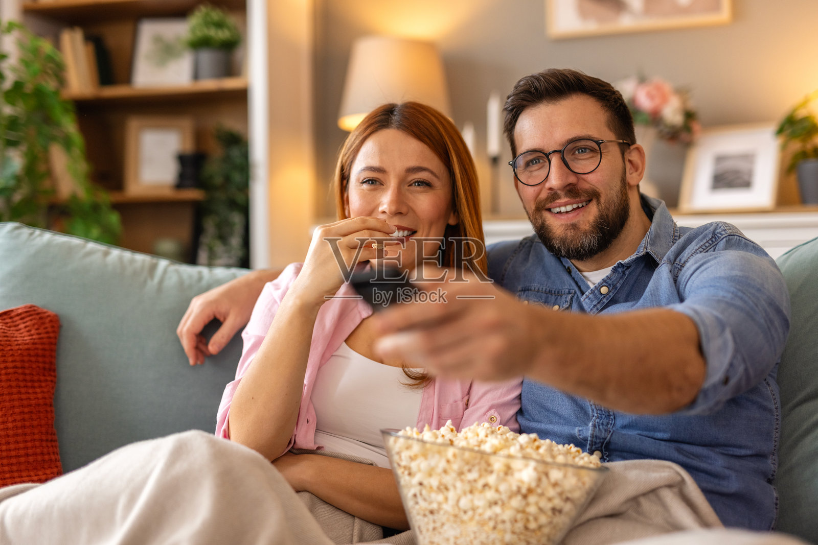 一对快乐的情侣在家享受温馨的电影之夜，坐在沙发上，手里拿着一碗爆米花。他们面带微笑，投入其中，共同度过一个有趣而放松的夜晚。照片摄影图片