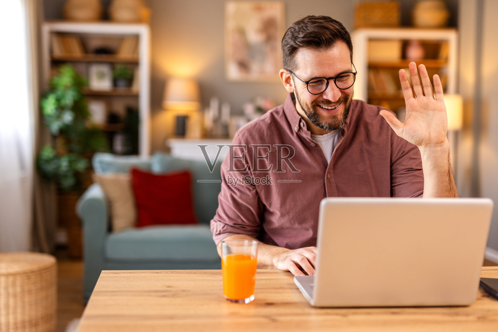 戴眼镜的年轻男子在视频通话中挥手，正在家中工作。他坐在明亮的厨房里，面前有一台笔记本电脑和一杯橙汁，享受着轻松而高效的氛围。照片摄影图片