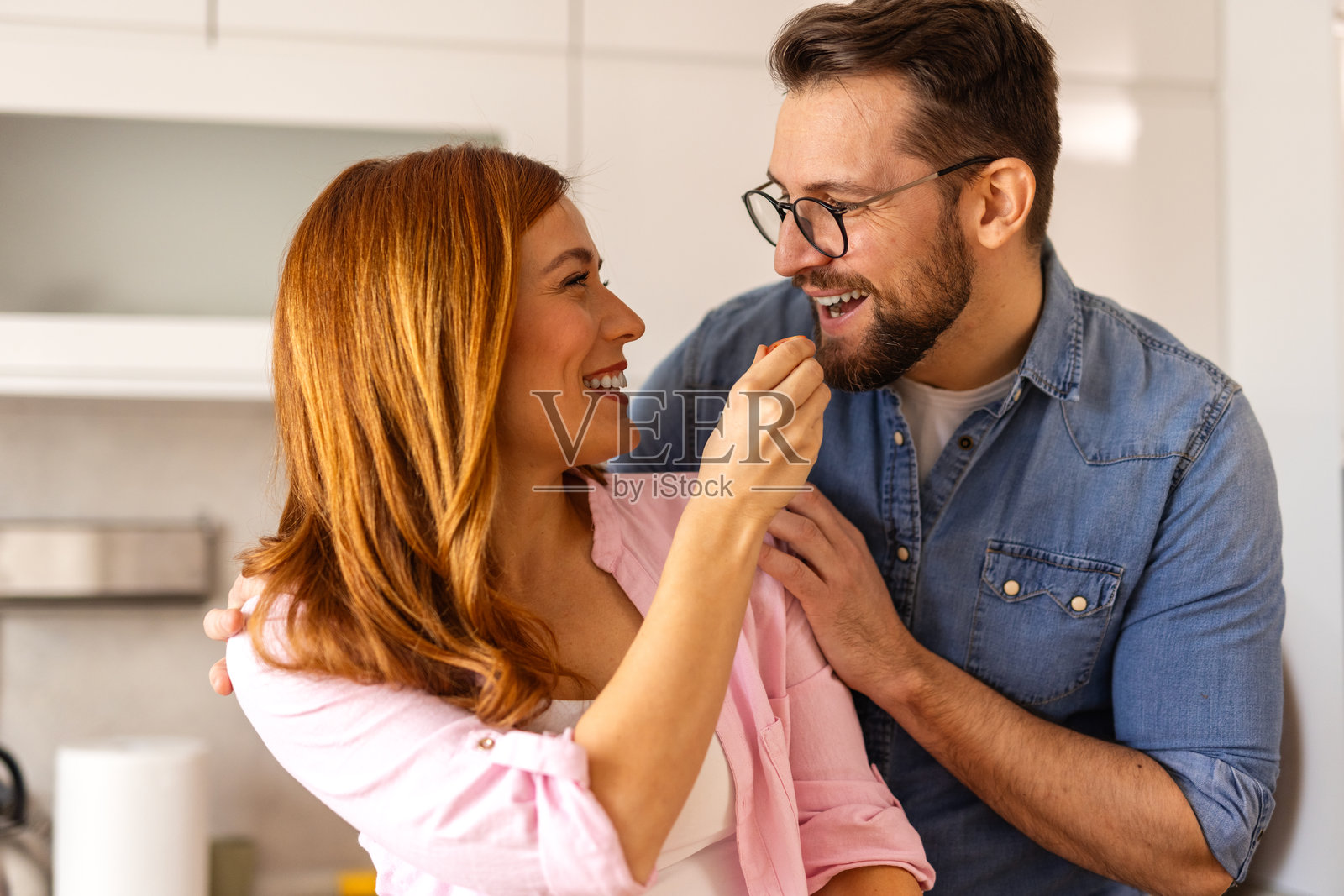 美丽的年轻情侣在家里的厨房里互相喂食并微笑。快乐的情侣正在明亮的厨房里准备健康的食物。健康饮食概念。照片摄影图片