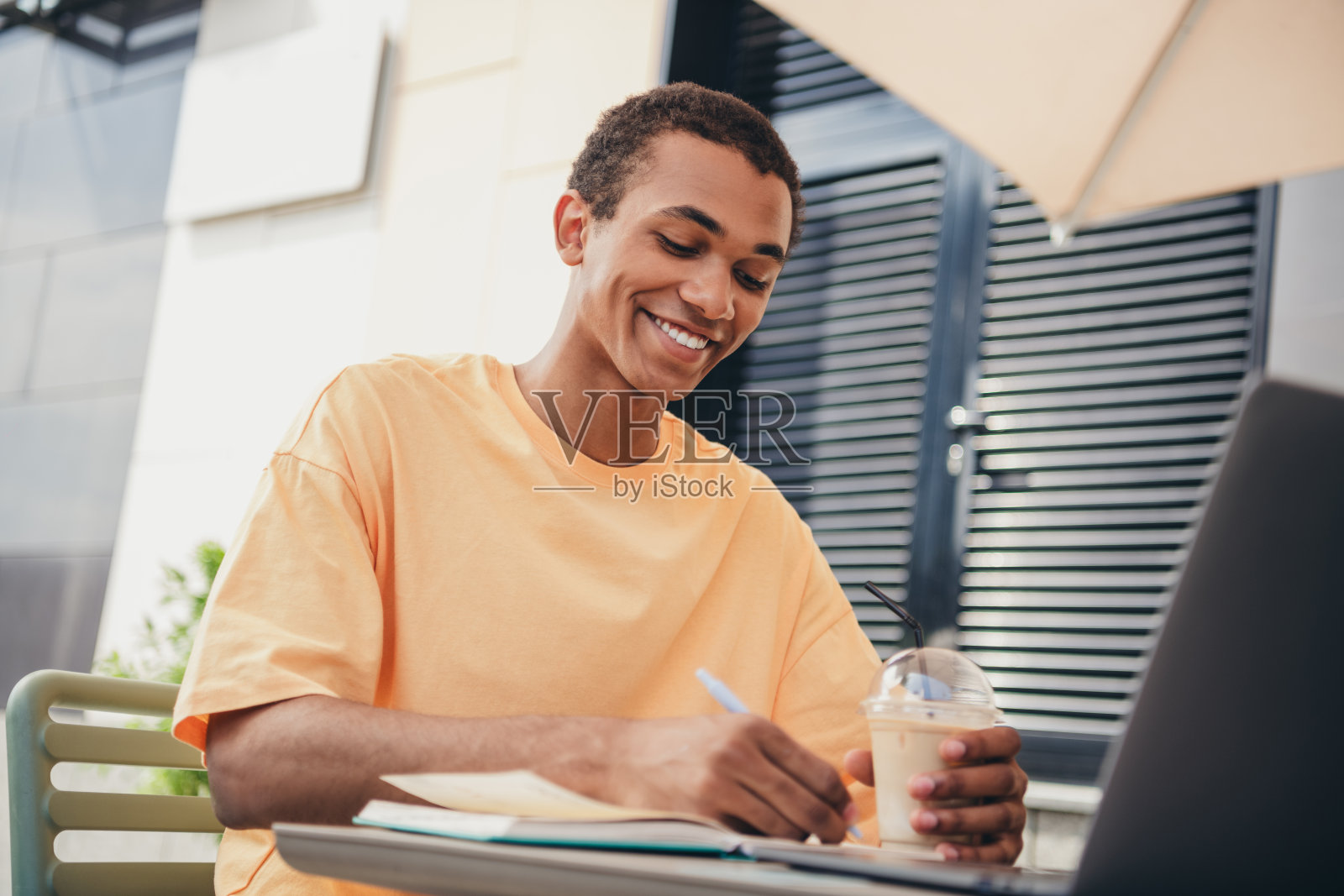 年轻男学生在阳光明媚的日子里，穿着时尚休闲的服装，享受户外学习时光，手边有冰咖啡，正在写笔记。照片摄影图片