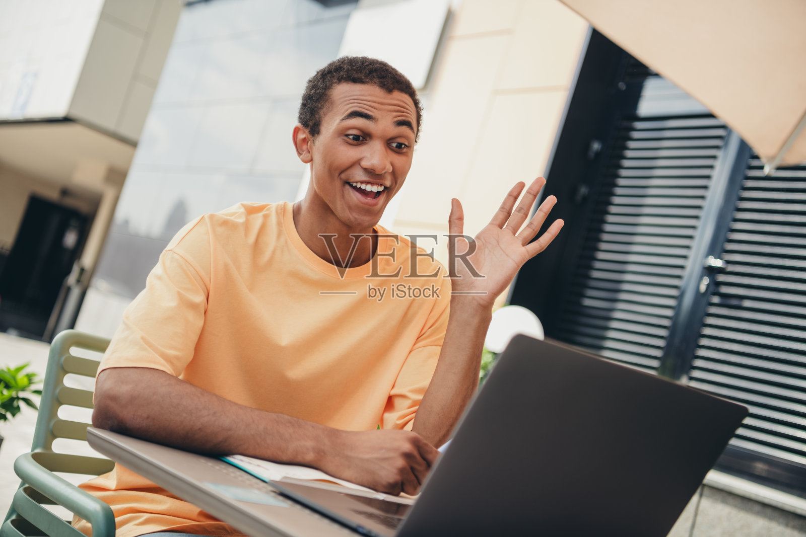 年轻的非裔美国男子在户外阳光明媚的日子里向笔记本电脑屏幕挥手，周围是城市氛围和休闲的环境。照片摄影图片